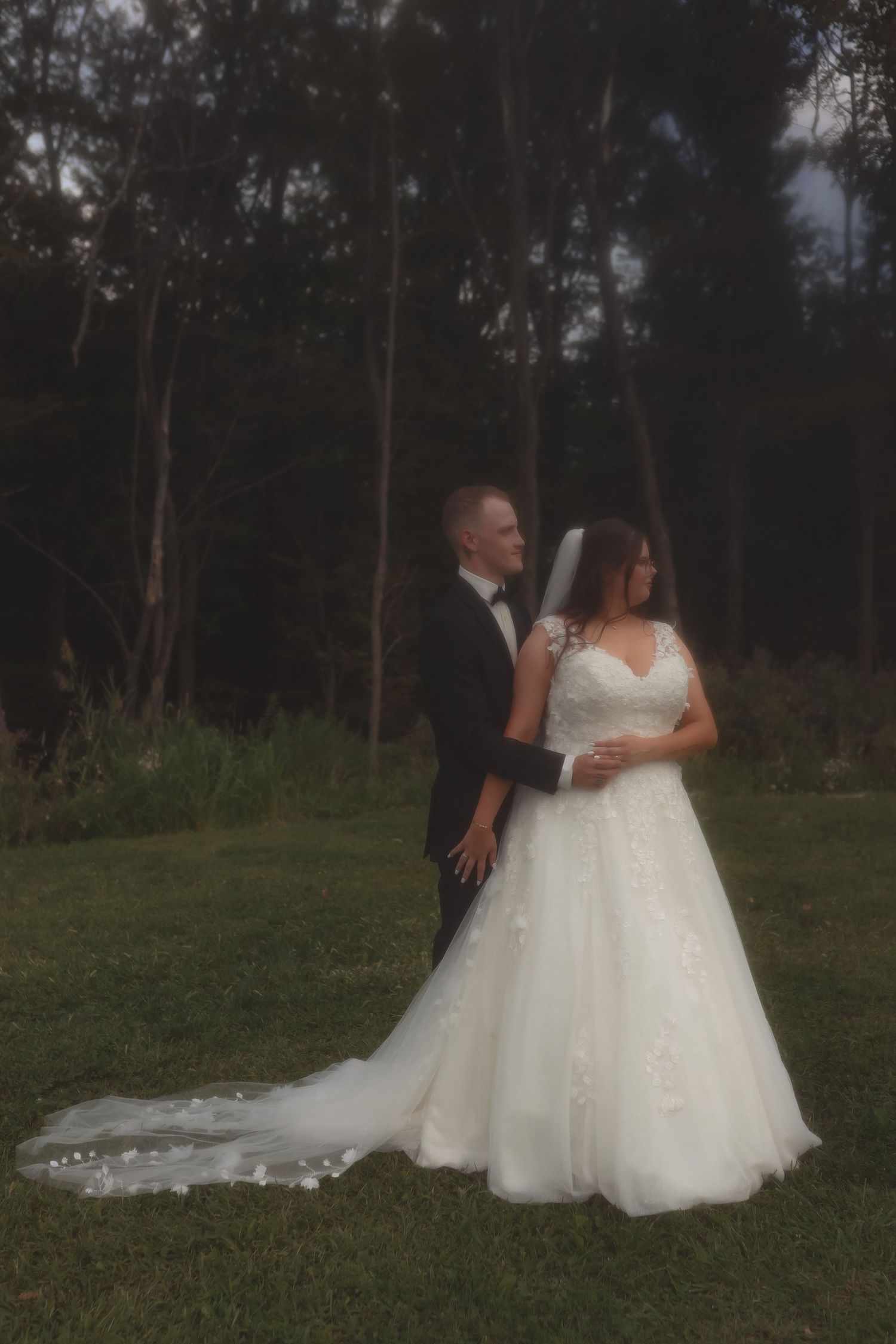 Bride and groom share a romantic moment outdoors, with the bride in a white ballgown and long veil among tall trees.