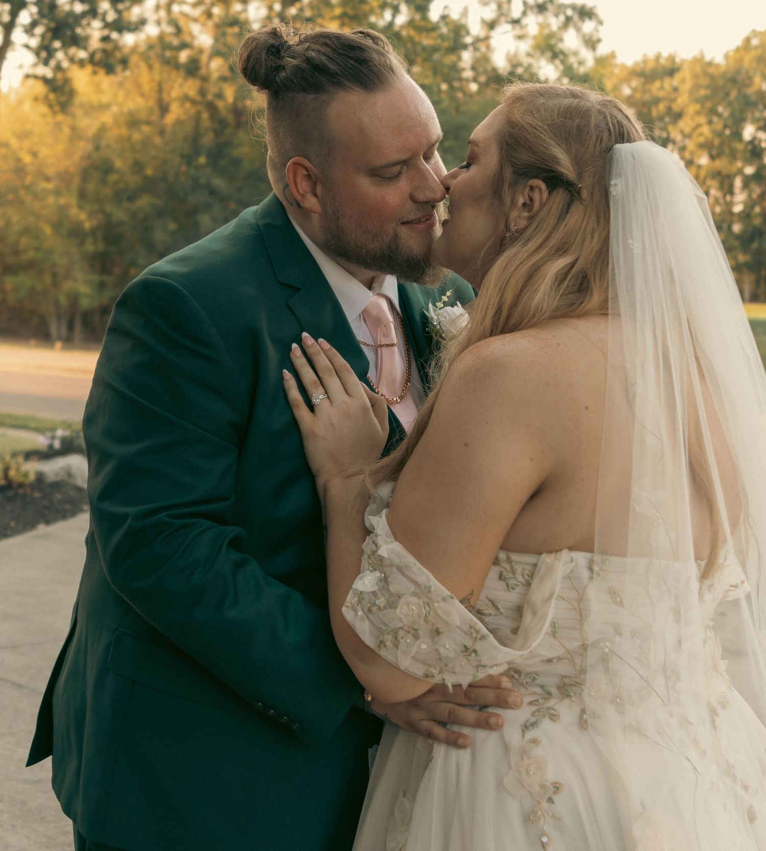 A couple shares a romantic kiss at their wedding, groom in green suit, bride in lace gown with veil.