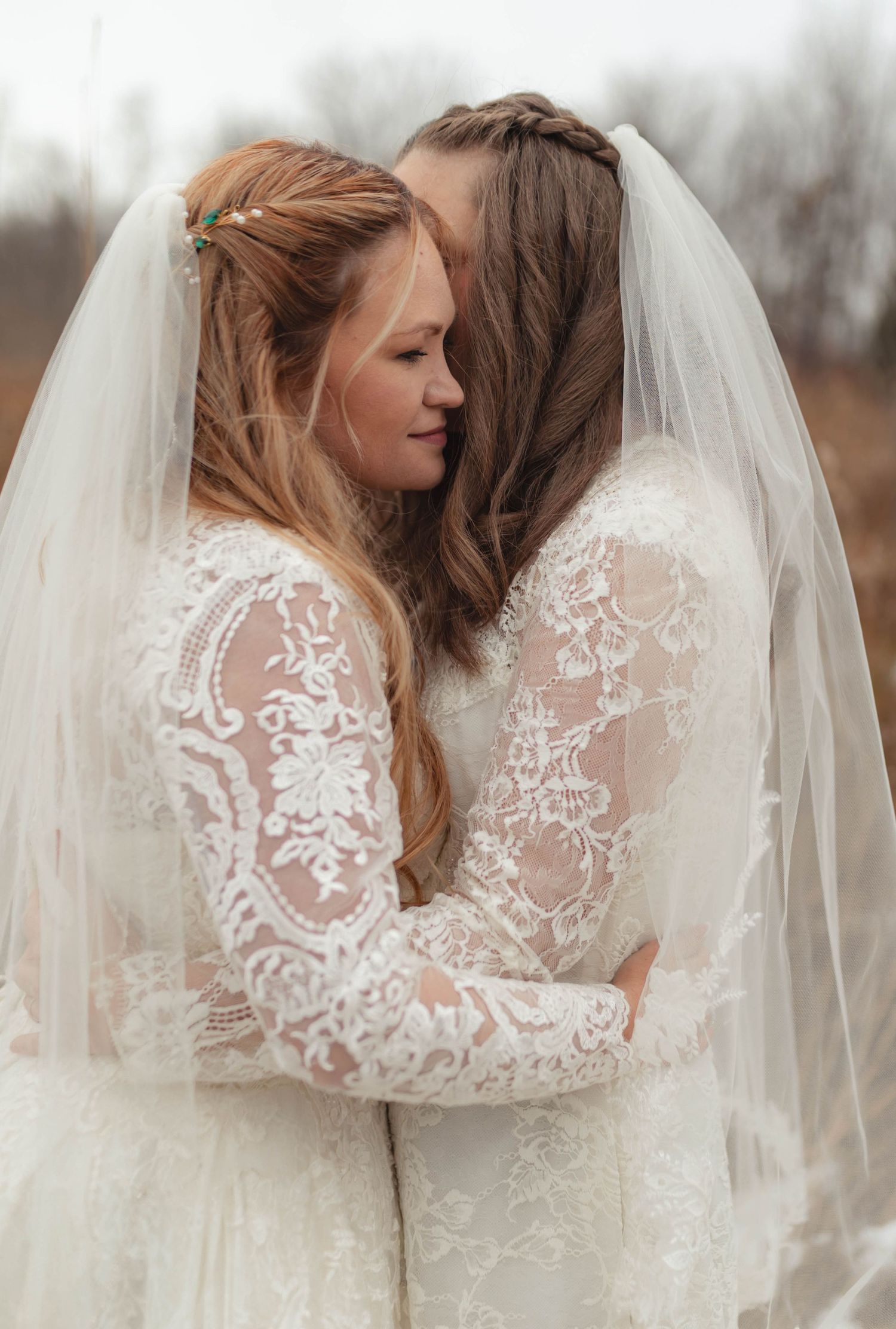 Two brides in lace wedding dresses share an intimate embrace under a veil outdoors.