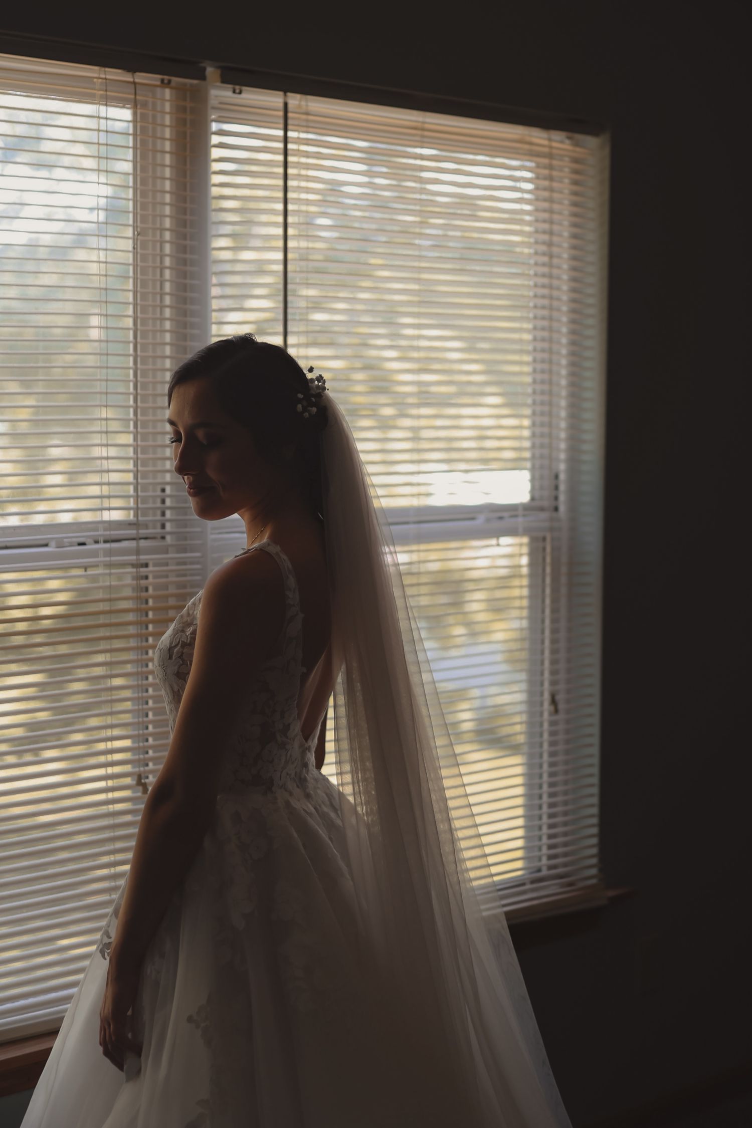 Bride in lace gown and veil stands in silhouette by window before wedding ceremony.