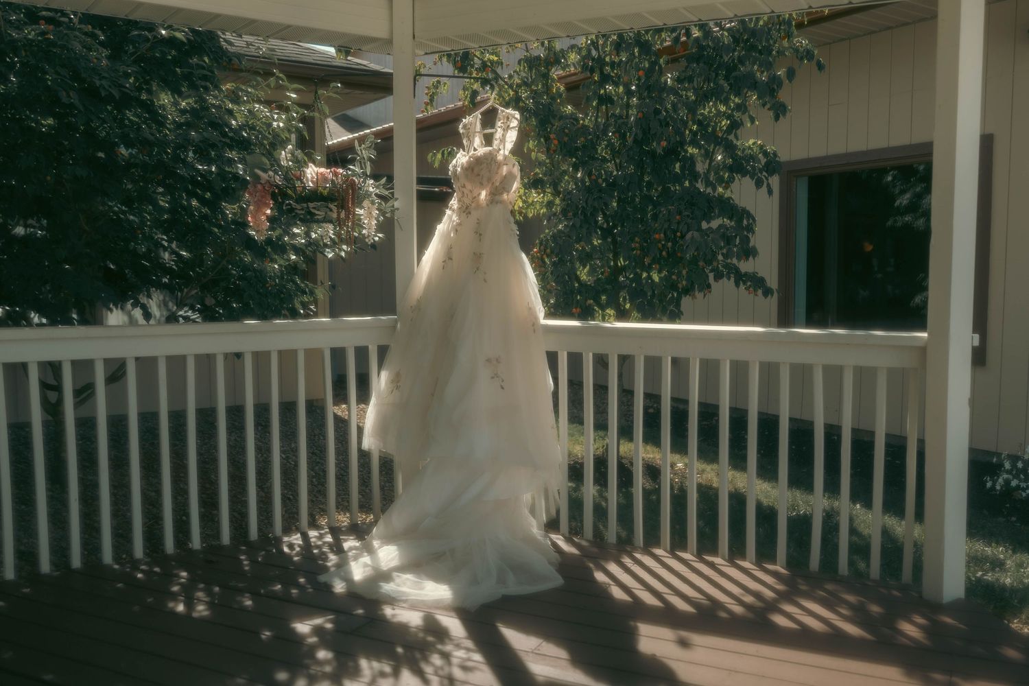 A white wedding dress hanging on a sunlit porch railing surrounded by greenery.