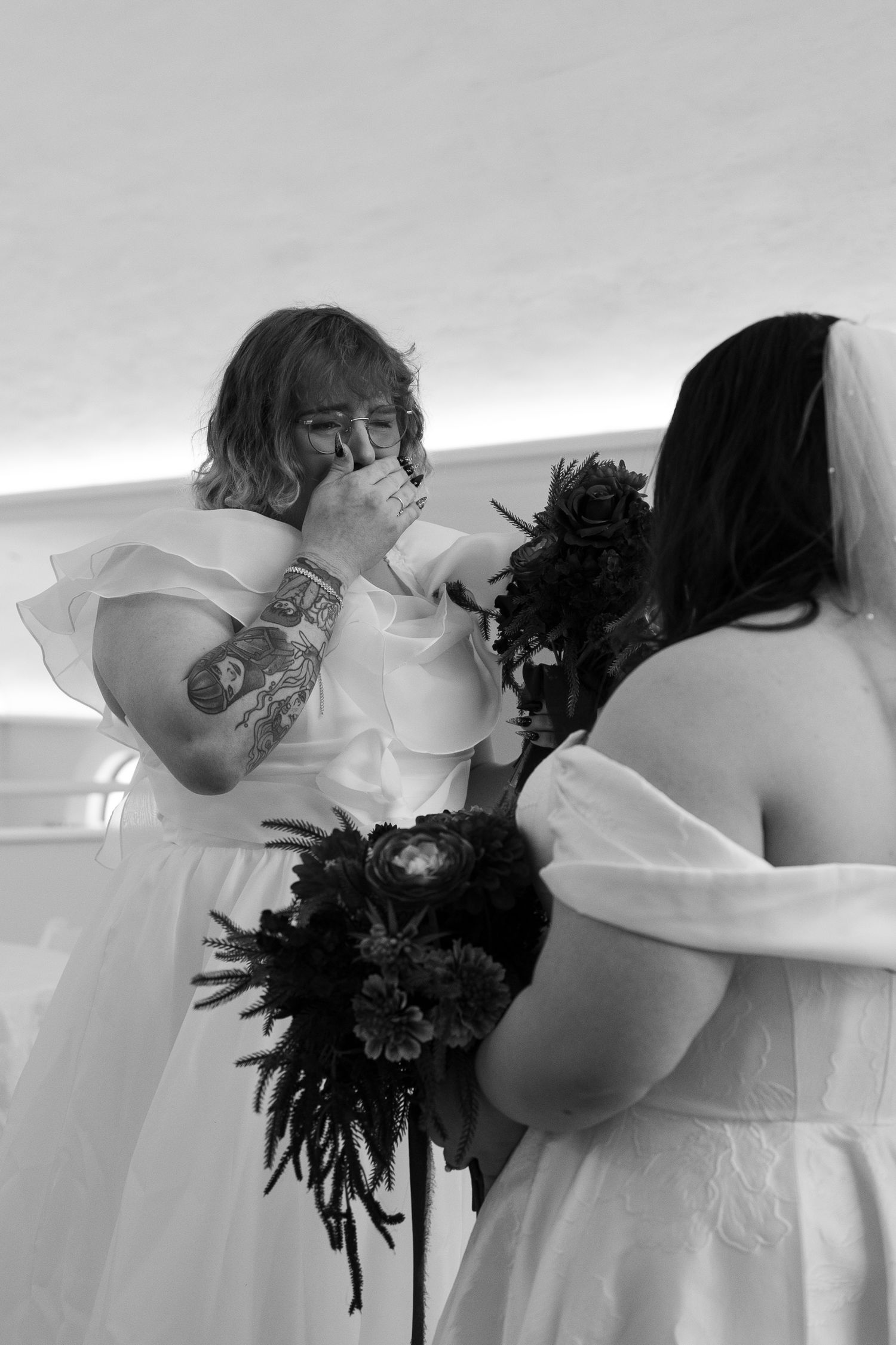 Two brides in white off-shoulder gowns share an emotional wedding moment, one wiping tears, holding dark floral bouquets.