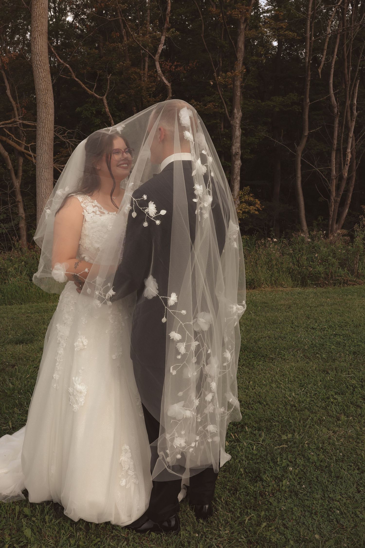 Smiling bride and groom share romantic moment under floral veil outdoors surrounded by lush greenery.