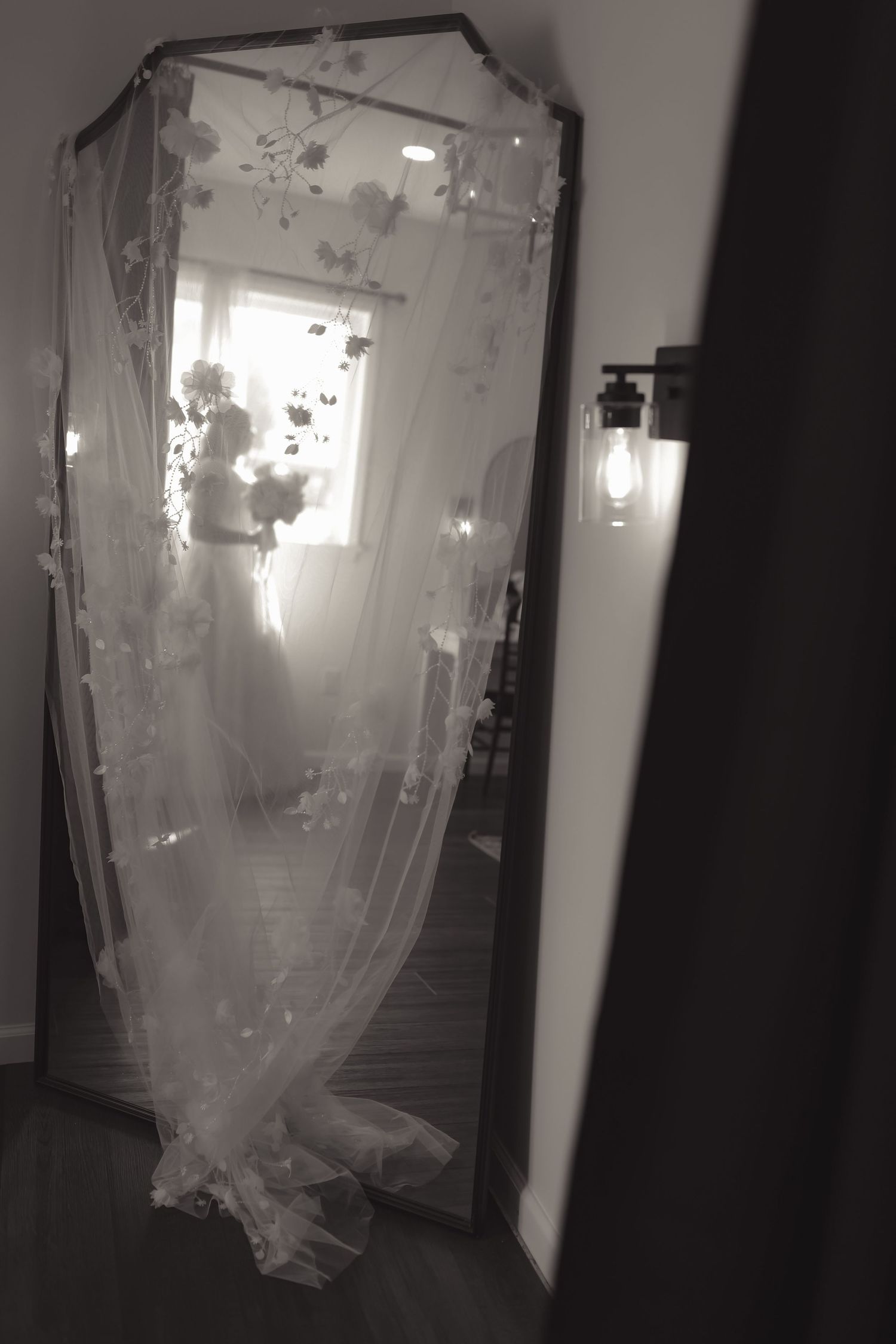 Black and white photo of a bride in a wedding dress reflected in a large floor mirror with floral veil.