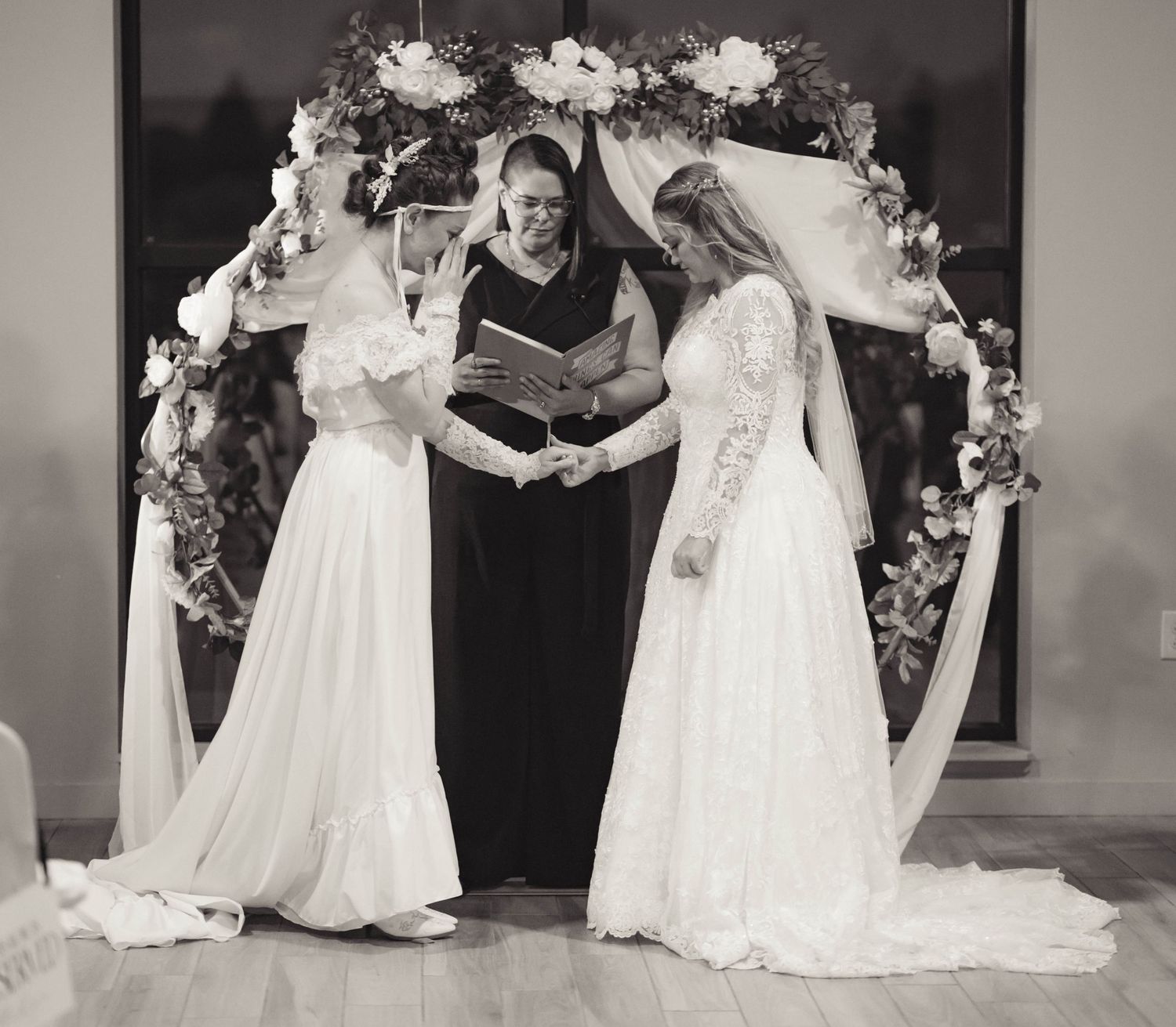 Black and white photo of two brides in white gowns exchanging vows under a floral arch with an officiant.