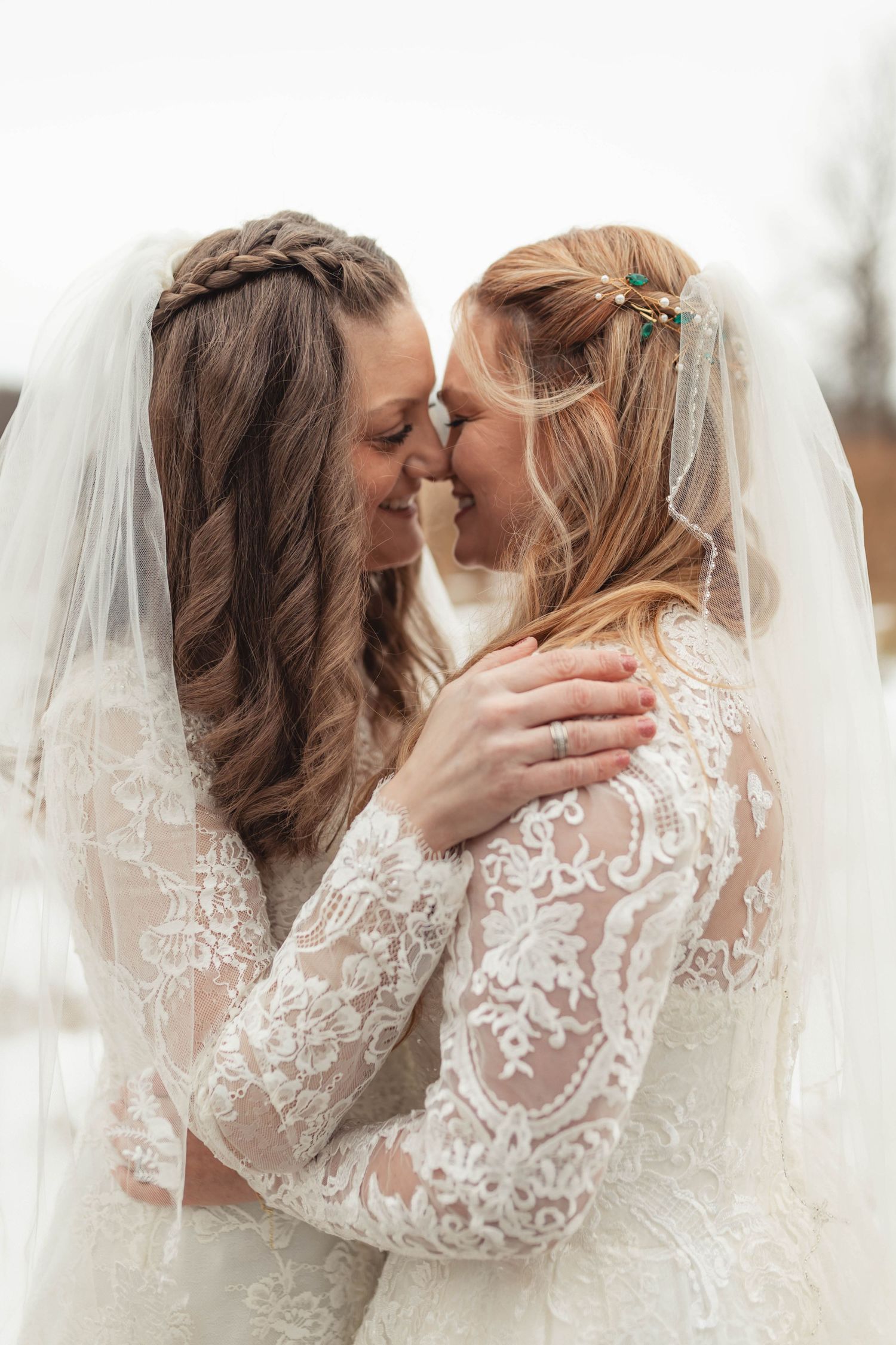 Two brides in white lace wedding dresses share an intimate moment under a veil, foreheads touching and smiling.