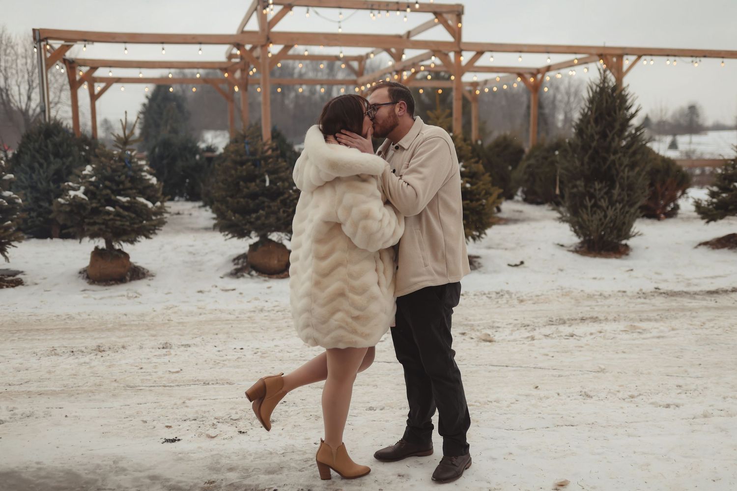 Couple embracing and kissing at a snowy Christmas tree farm with a wooden pergola backdrop.