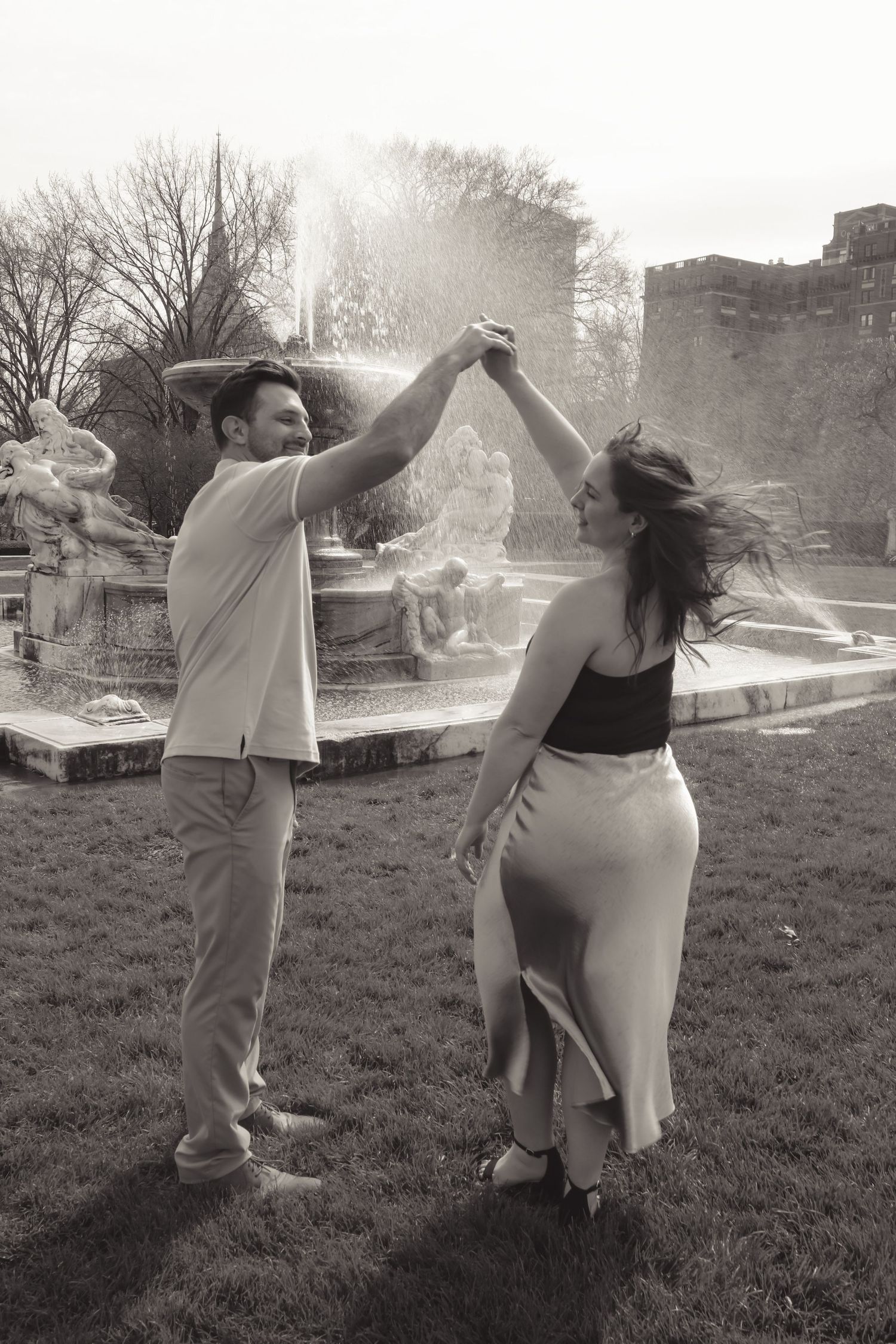 Black and white photo of a couple dancing joyfully in front of a grand fountain in a lush park.