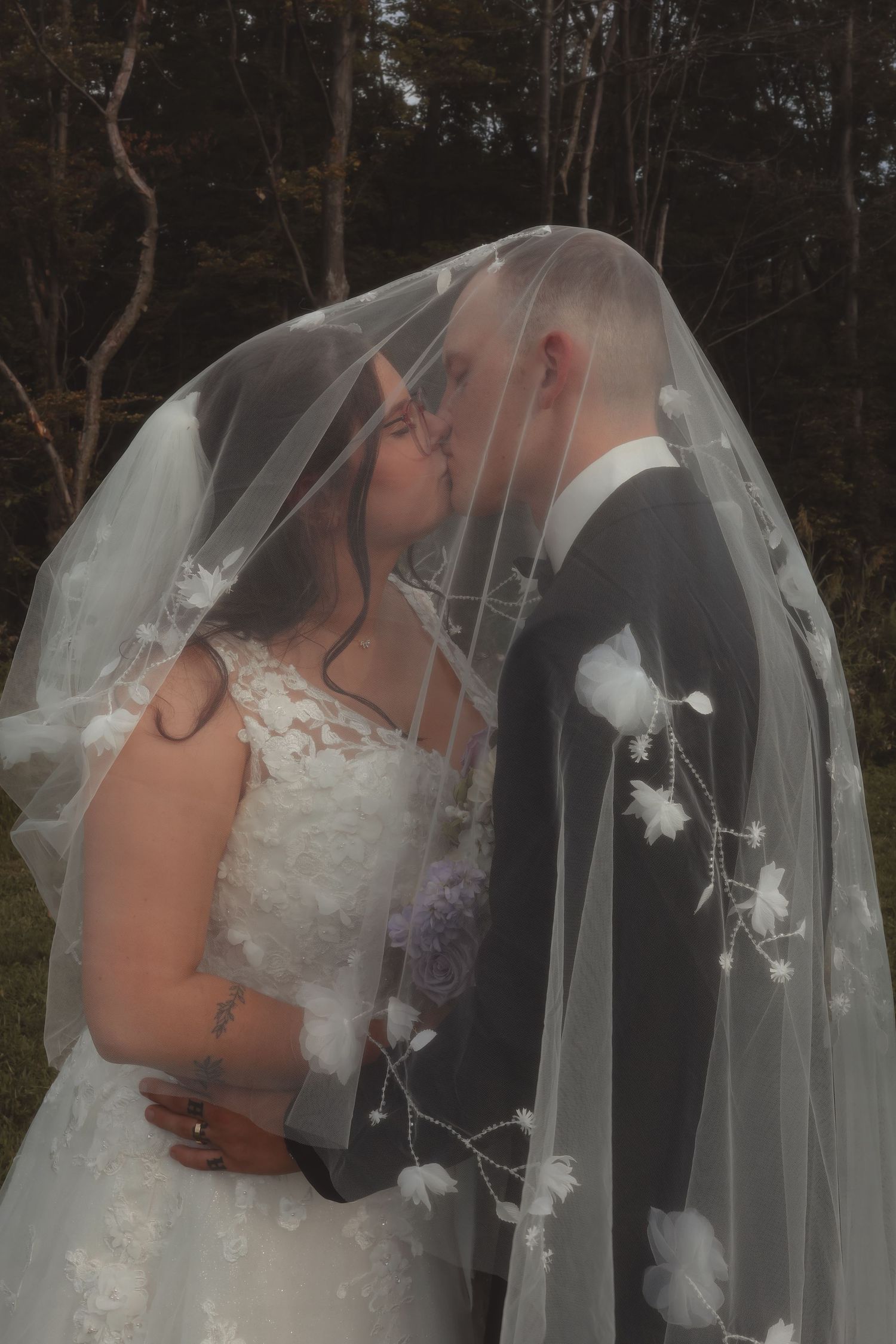 Bride and groom share a romantic kiss under a floral embroidered veil outdoors on their wedding day.