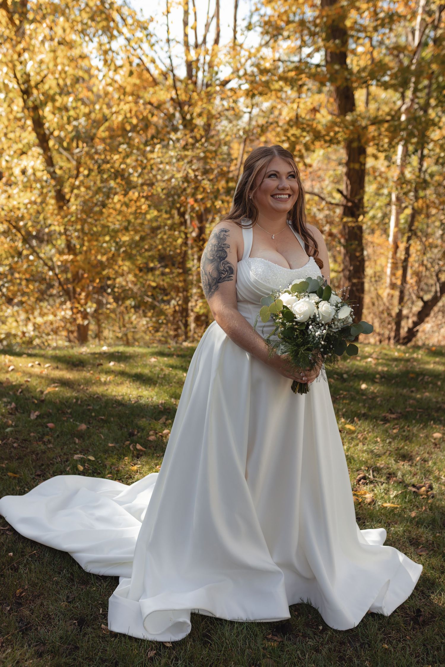 Bride in white ball gown with train holding bouquet among colorful autumn trees outdoors.