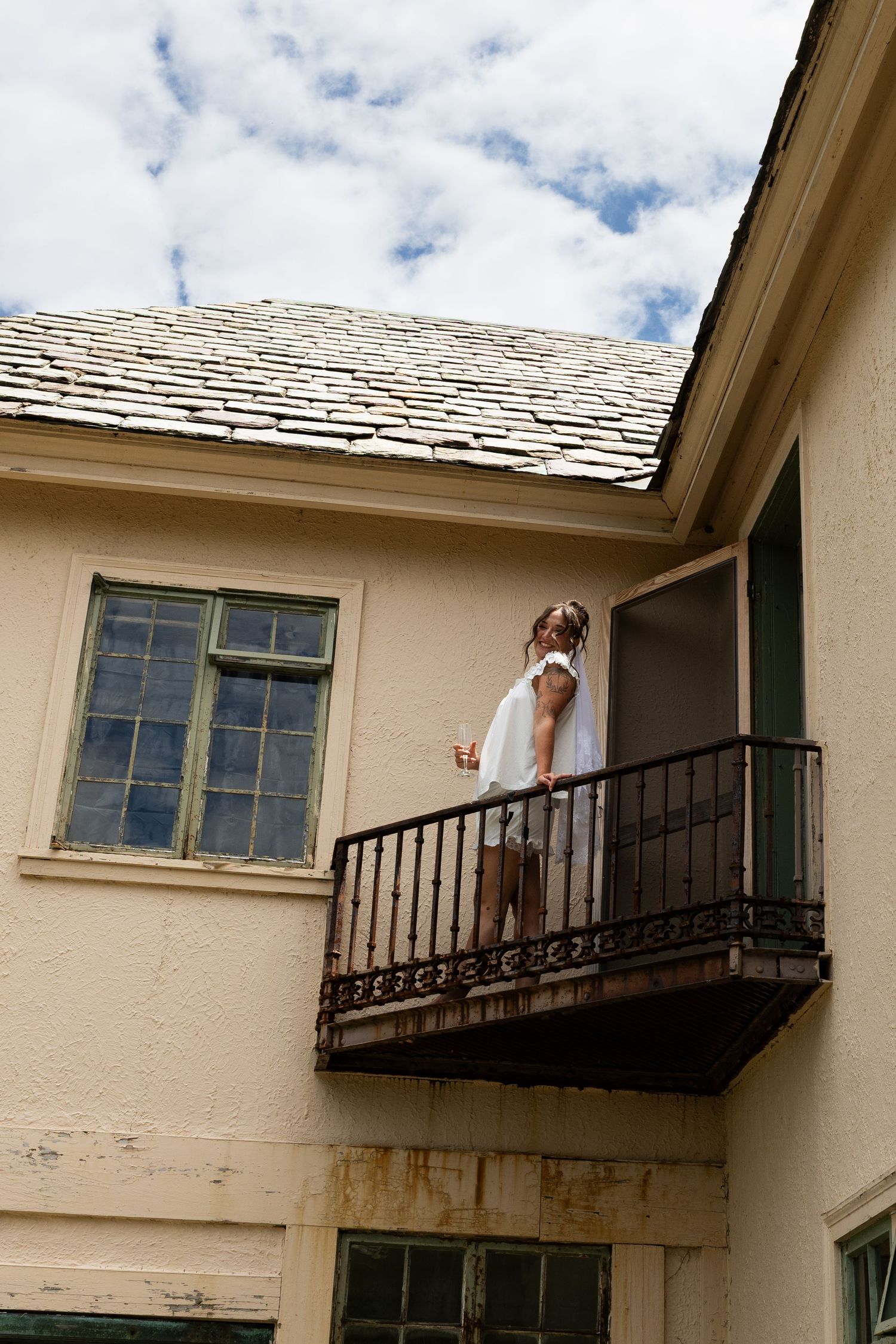 Woman in white dress standing on a rustic iron balcony of a vintage beige building under a cloudy sky.
