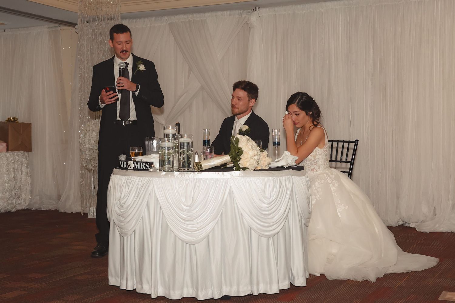 Best man giving a wedding toast at a reception while bride and groom sit at a decorated head table.