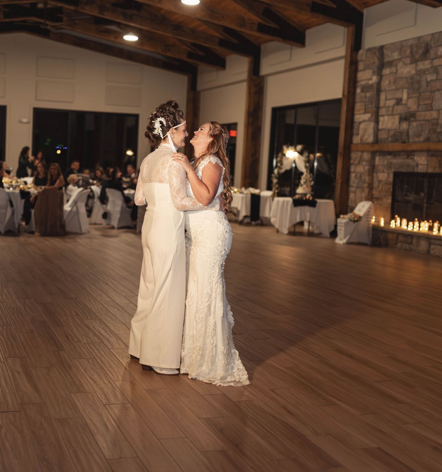 Two brides share their first dance at a rustic wedding reception venue with warm lighting and stone fireplace.