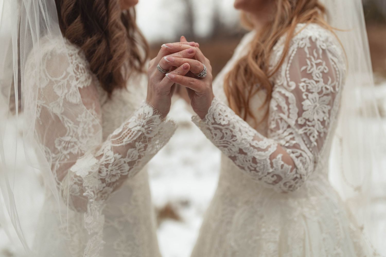 Two brides in lace wedding gowns holding hands outdoors in a snowy winter setting.