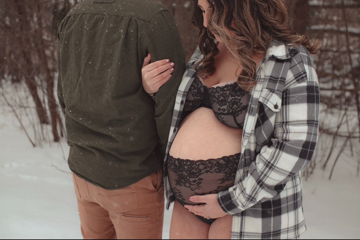 A maternity photo in winter showing a plaid flannel shirt and olive sweater against snowy background.