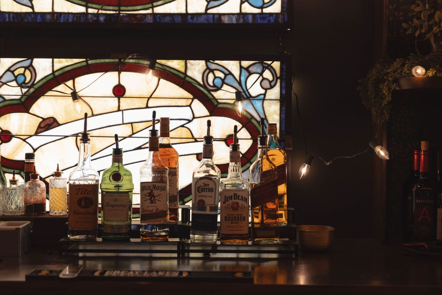 Bar shelf with whiskey bottles displayed in front of a colorful stained glass window with string lights.