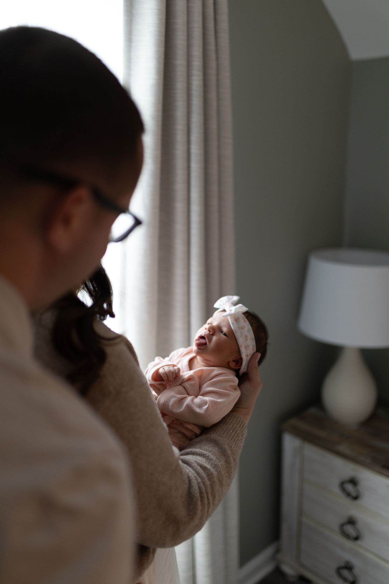 A mother in glasses holds her newborn baby wearing a white bow headband near a window in a softly lit nursery.