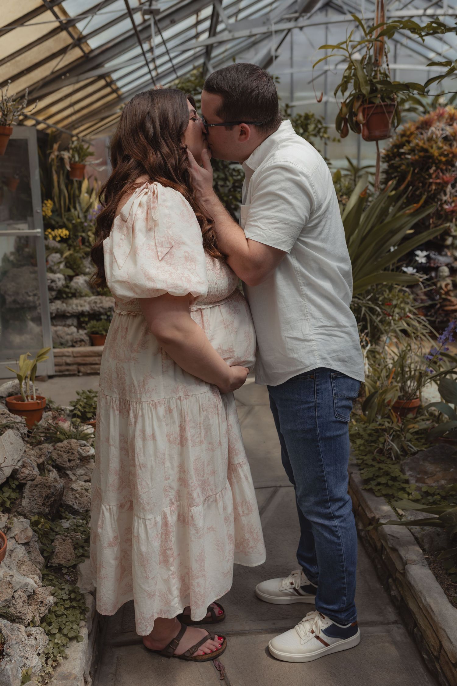 Pregnant woman in floral dress kissed by partner in greenhouse surrounded by lush tropical plants.