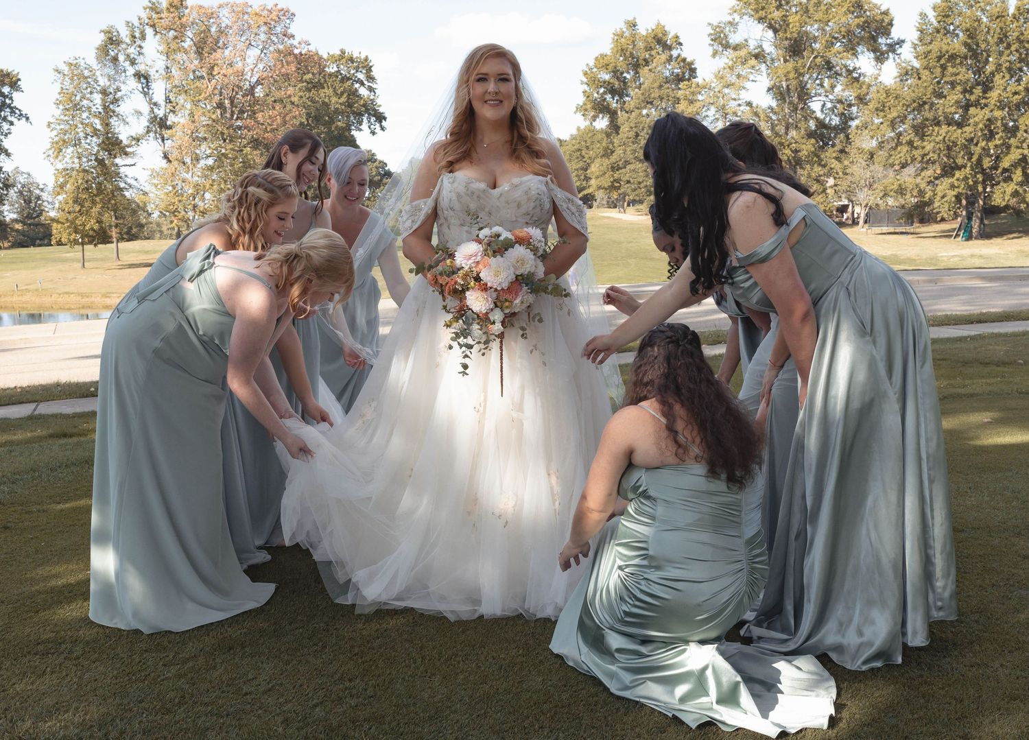 Bride in a ball gown surrounded by bridesmaids in sage green dresses adjusting her train outdoors.