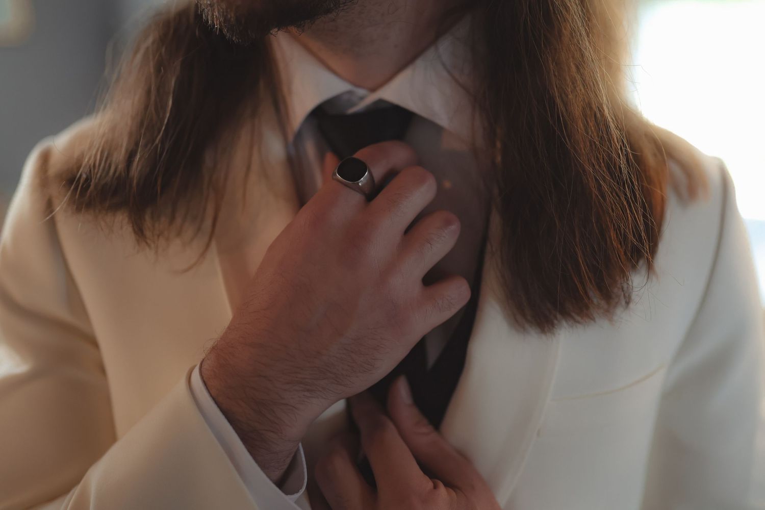 A groom in a white suit adjusts his dark tie, wearing a black ring, in a close-up wedding preparation shot.