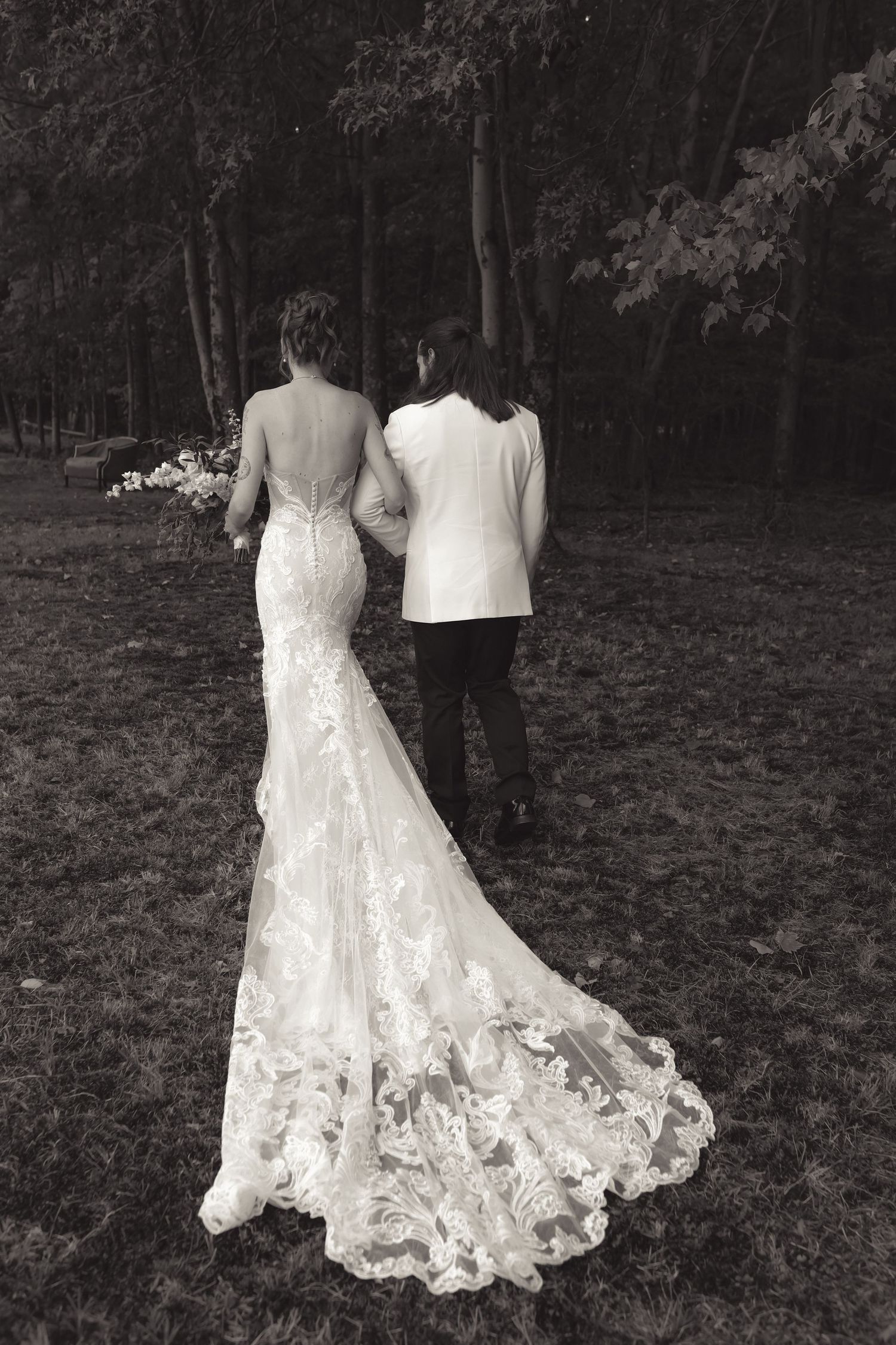 Black and white photo of bride in lace gown with train walking arm in arm with groom through wooded area.