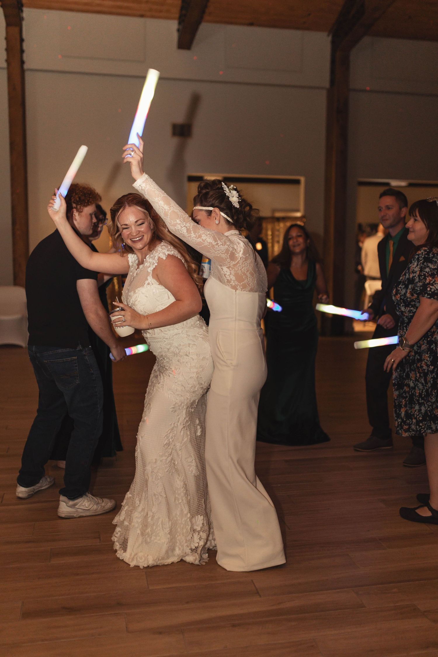 Two brides in lace wedding dresses dance with glowsticks at their wedding reception.