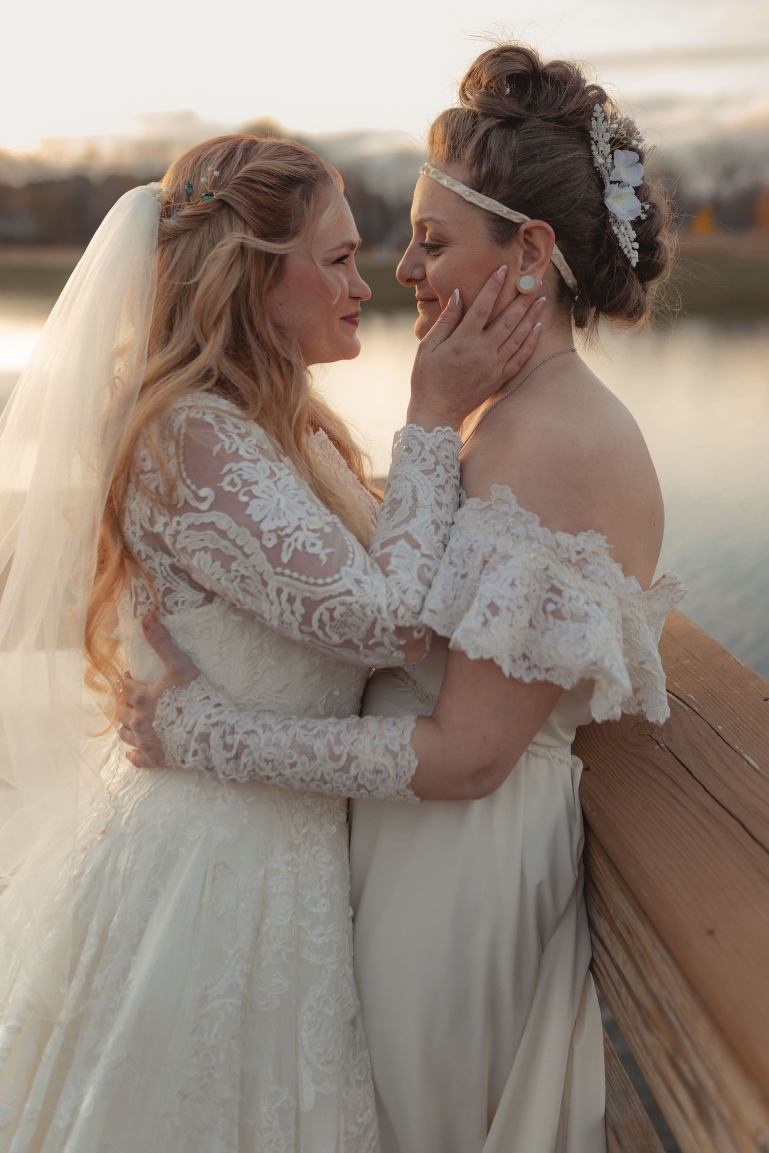 Two brides in lace wedding gowns embracing tenderly near a lake at sunset.