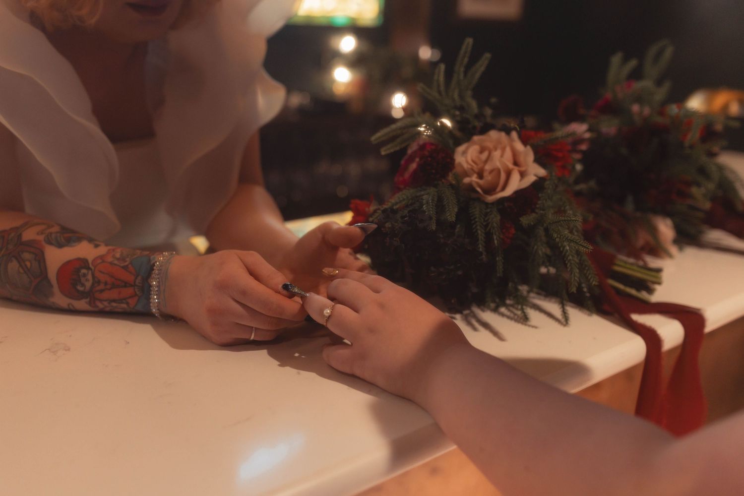 Tattooed bride placing wedding ring on groom's finger, with a floral bouquet featuring roses and greenery nearby.