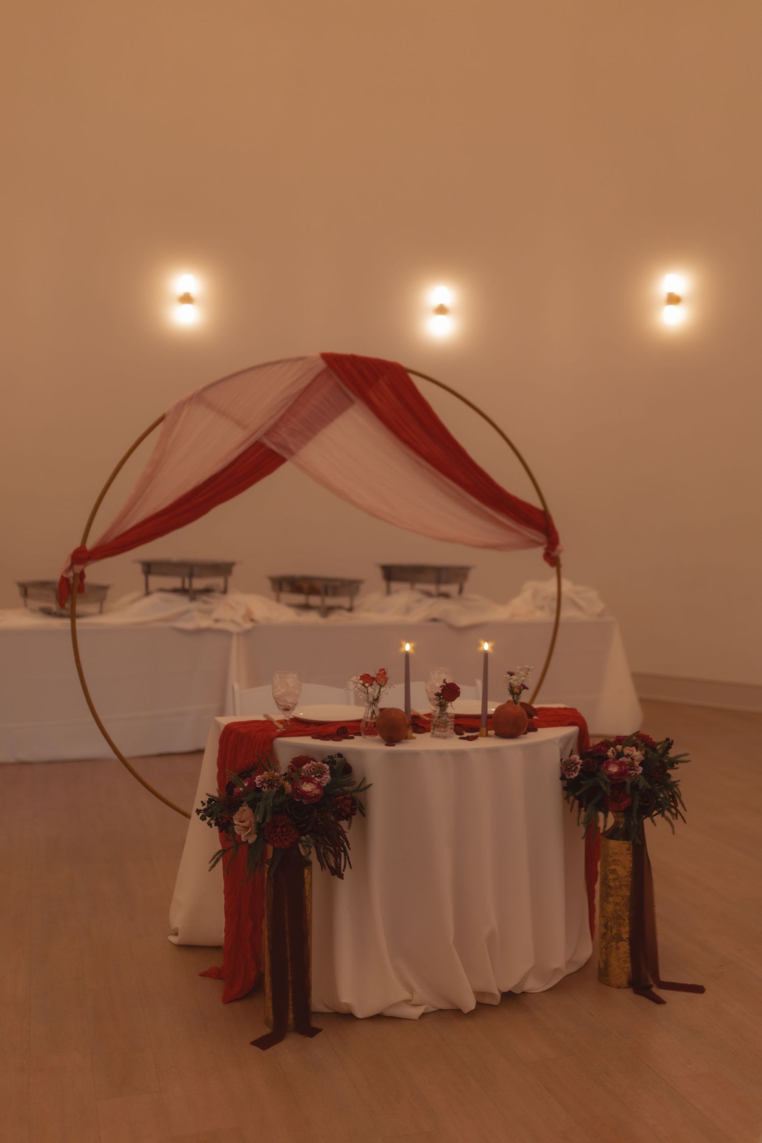 Elegant sweetheart table with circular gold arch, red draping, candles, and floral arrangements for a wedding reception.