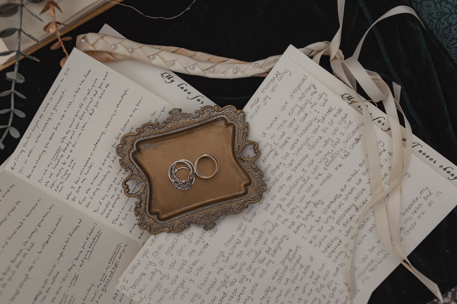 Two gold wedding rings rest on a vintage ornate tray atop handwritten love letters and wedding vows.