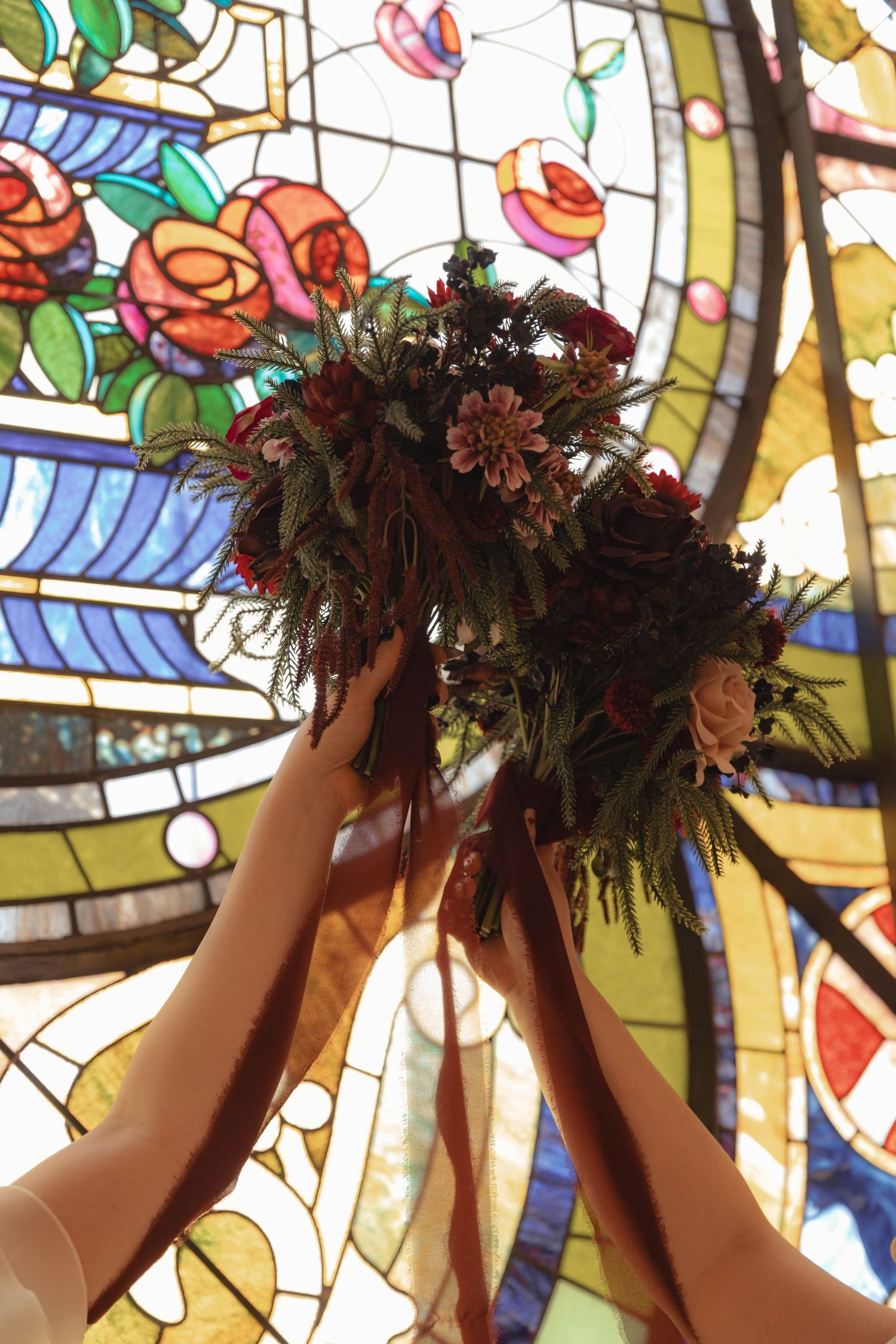 Hands raise a lush dark floral bouquet against a colorful stained glass church window.