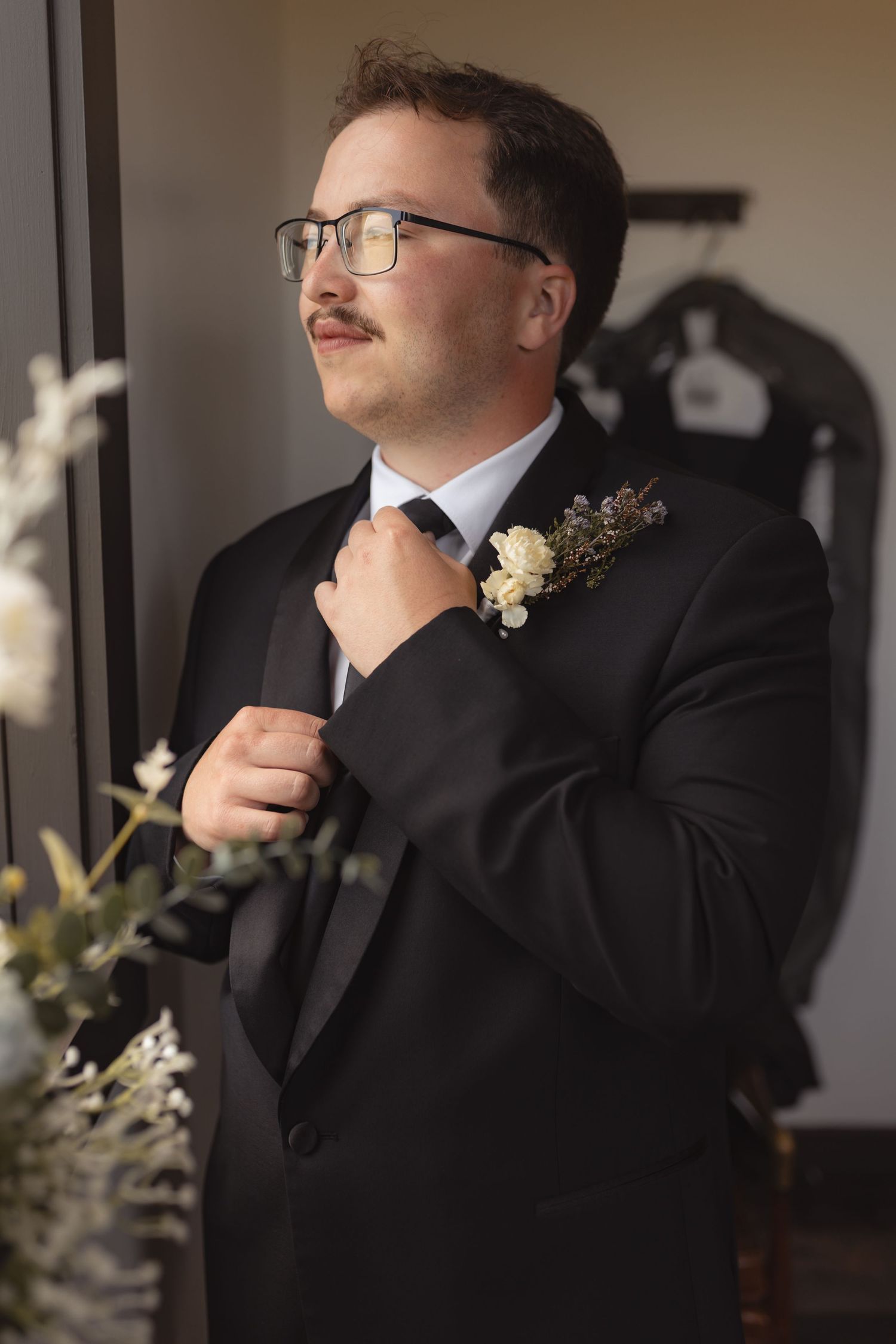 A groom in a black tuxedo adjusts his tie while wearing glasses and a floral boutonniere, preparing for his wedding day.