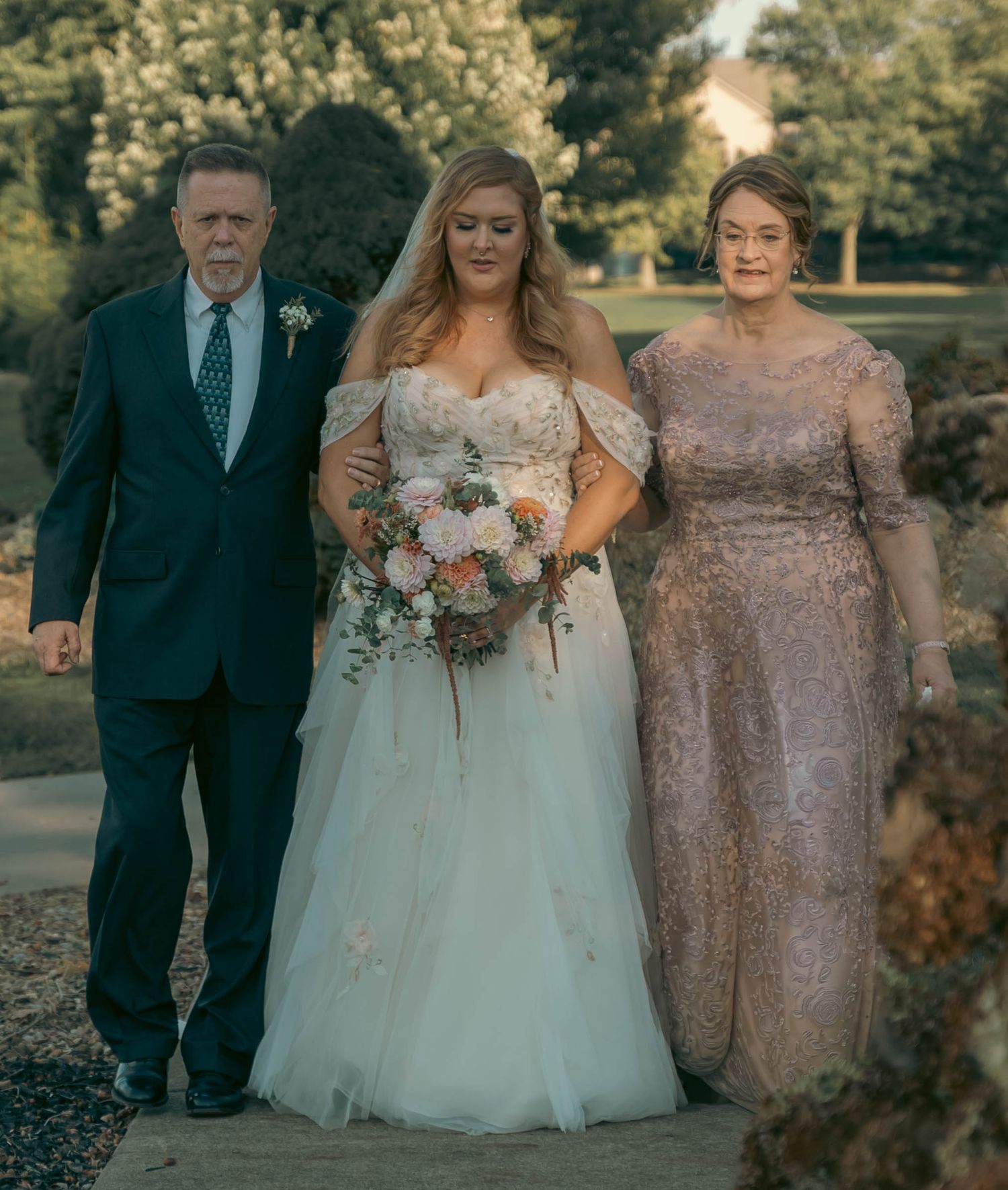 Bride in white gown holding bouquet walks with parents in formal attire at outdoor wedding ceremony.