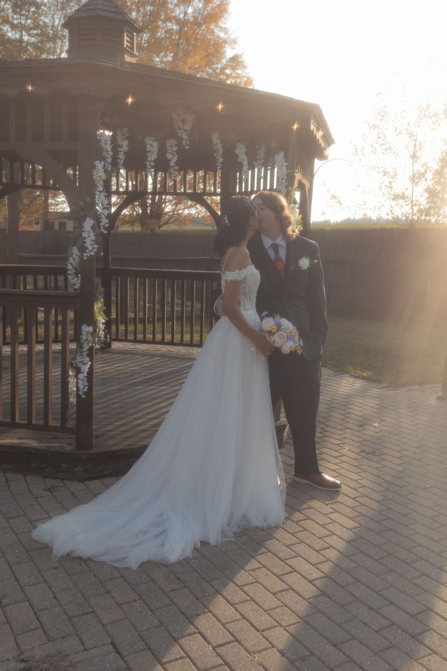 Bride and groom sharing romantic moment near decorated gazebo in golden sunset light outdoors.