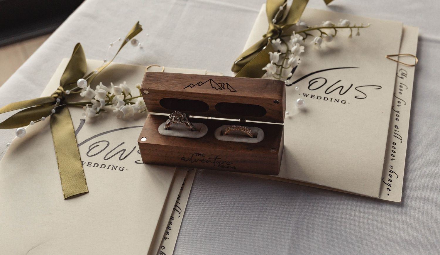 Elegant wooden ring box with wedding rings displayed on OWS Wedding stationery, adorned with delicate white flowers.