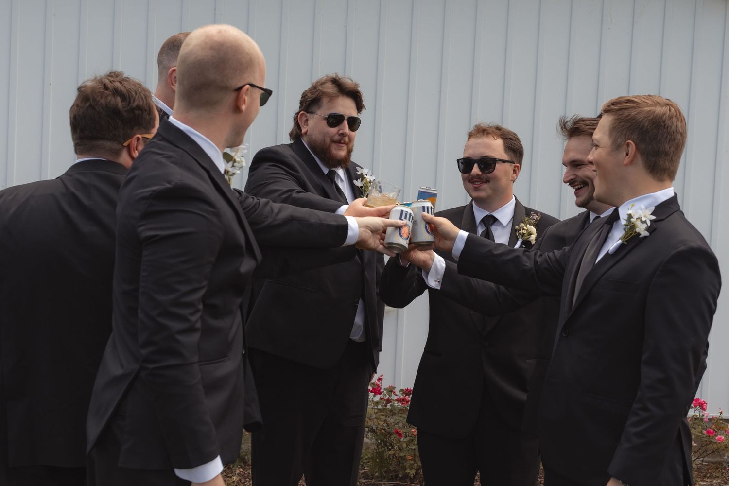 Groomsmen in black suits toasting together outdoors at a wedding celebration.