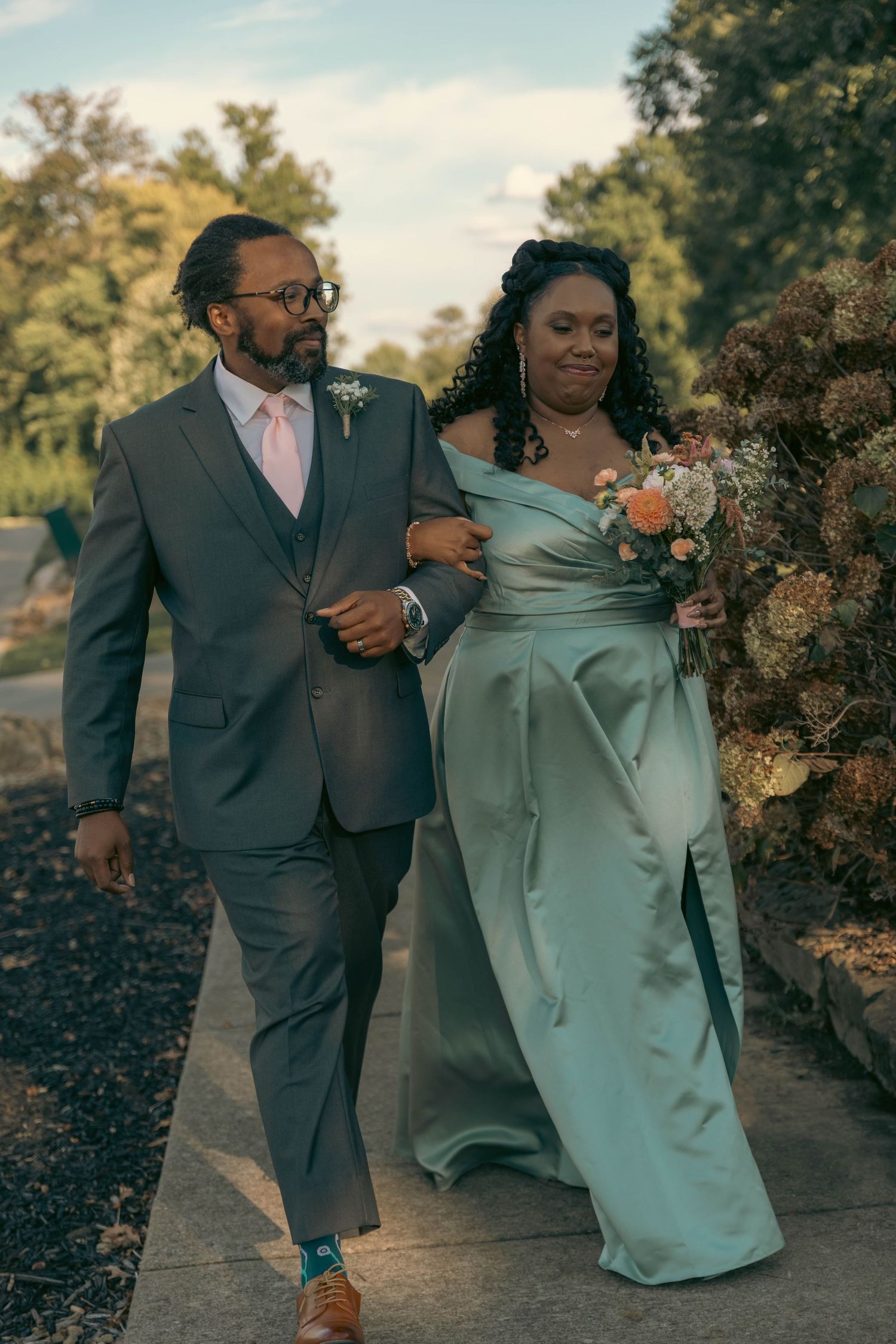 Elegant couple walks arm-in-arm, groom in gray suit, bride in sage green gown holding floral bouquet outdoors.