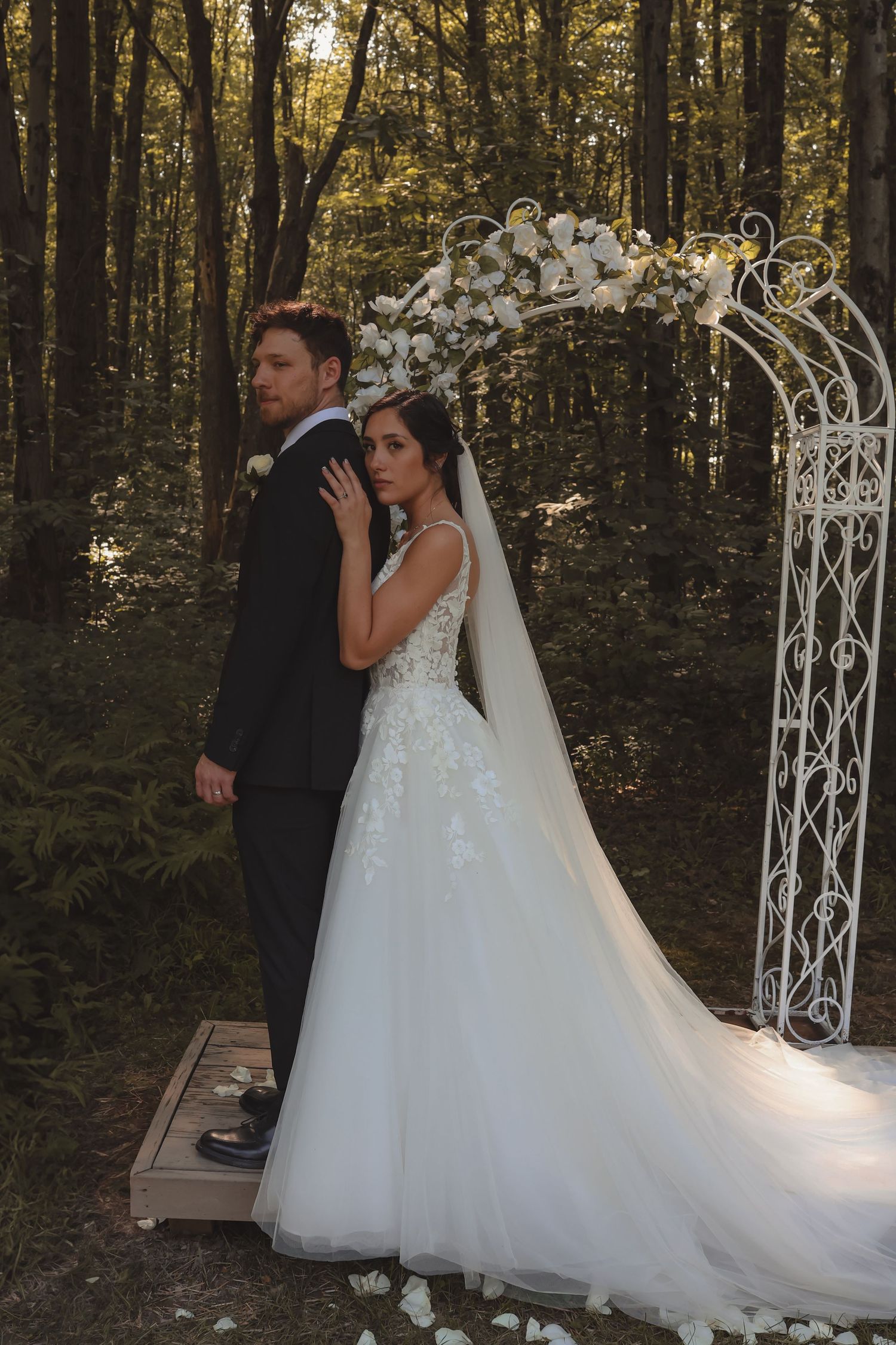 Bride in lace gown and groom in black suit stand back-to-back at a white floral arch in a wooded setting.