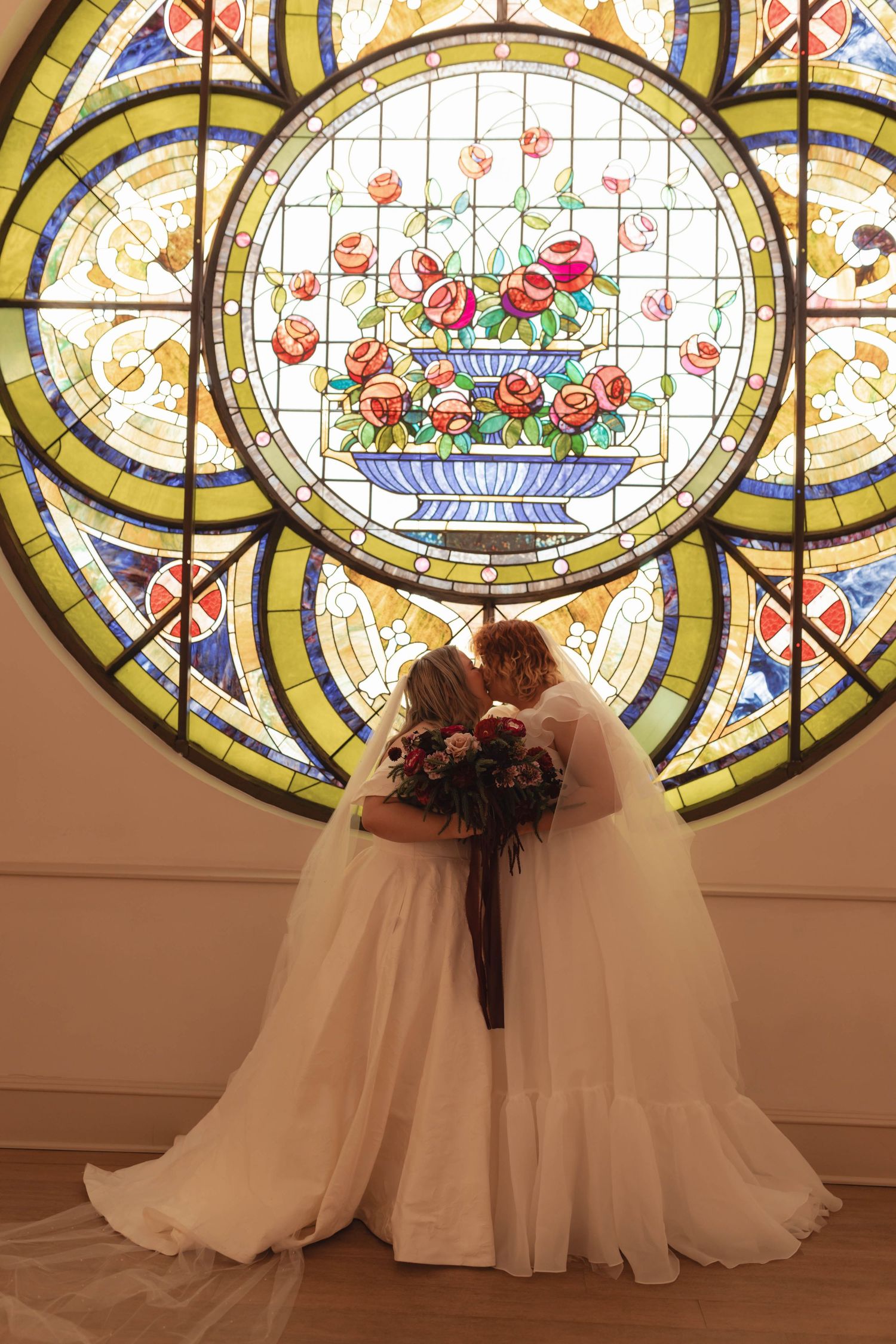 Two brides in white gowns share a kiss before a stunning circular stained glass window featuring a floral bouquet design.