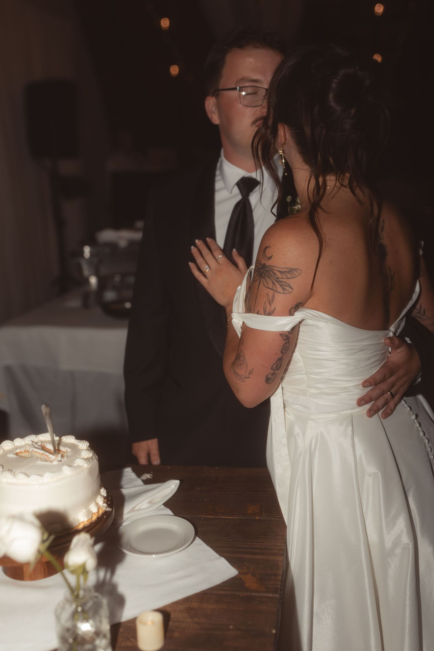 Bride and groom cutting their wedding cake, bride wearing white off-shoulder gown with tattoos, groom in black suit.