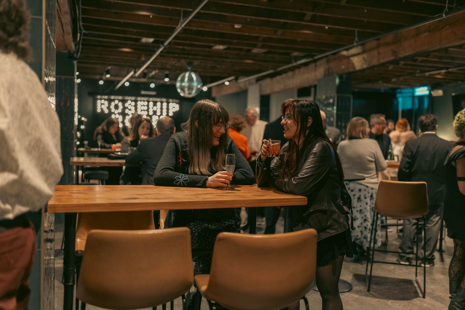 Two women chat over drinks at Rosehip bar, surrounded by patrons in a warmly lit, modern restaurant setting.