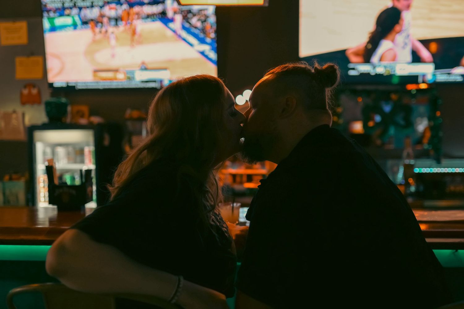 Couple sharing a romantic kiss at a dimly lit sports bar with TVs glowing in the background.