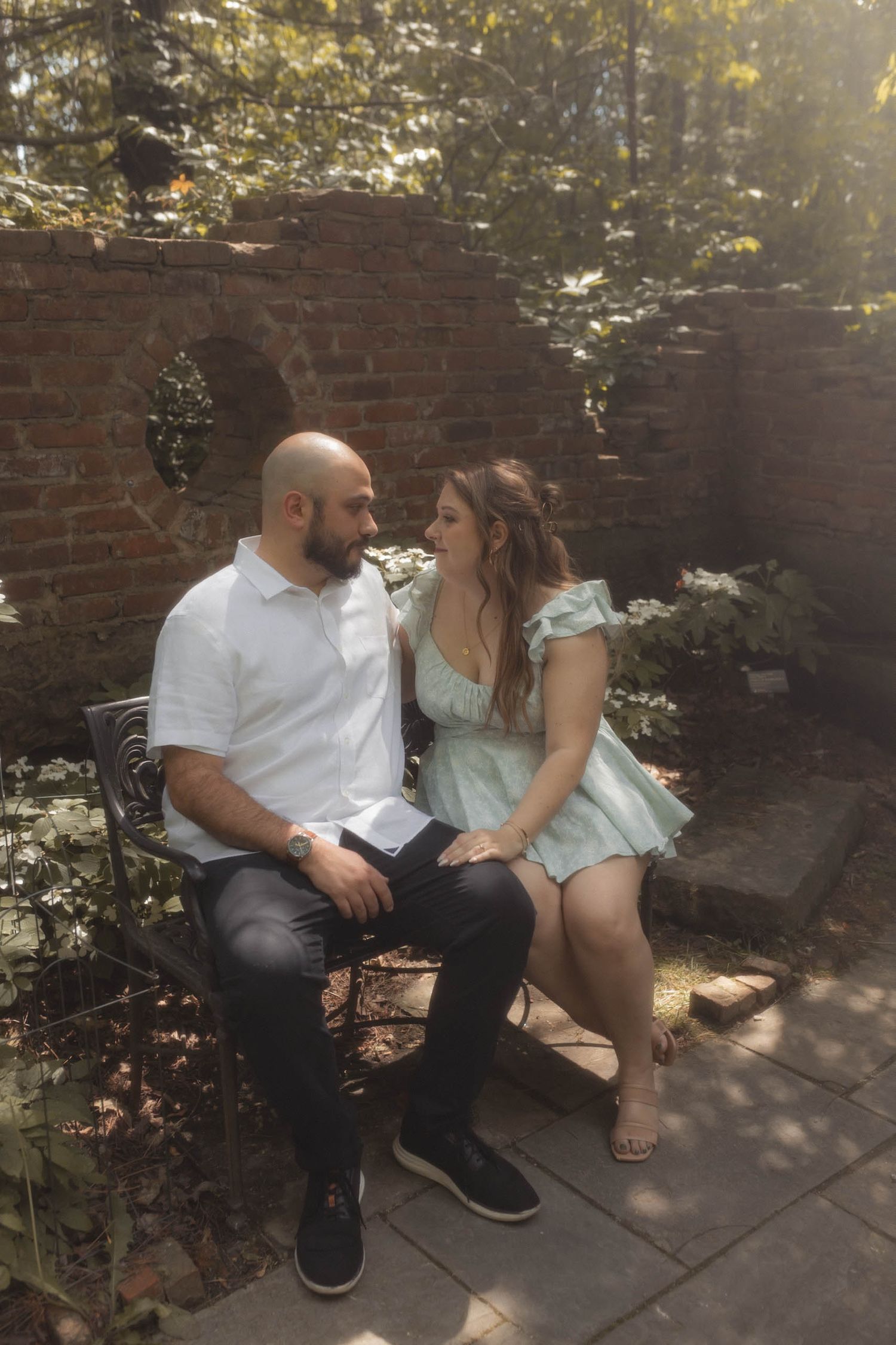 Couple sitting closely on a bench in a sunlit garden, gazing lovingly at each other near a brick wall.