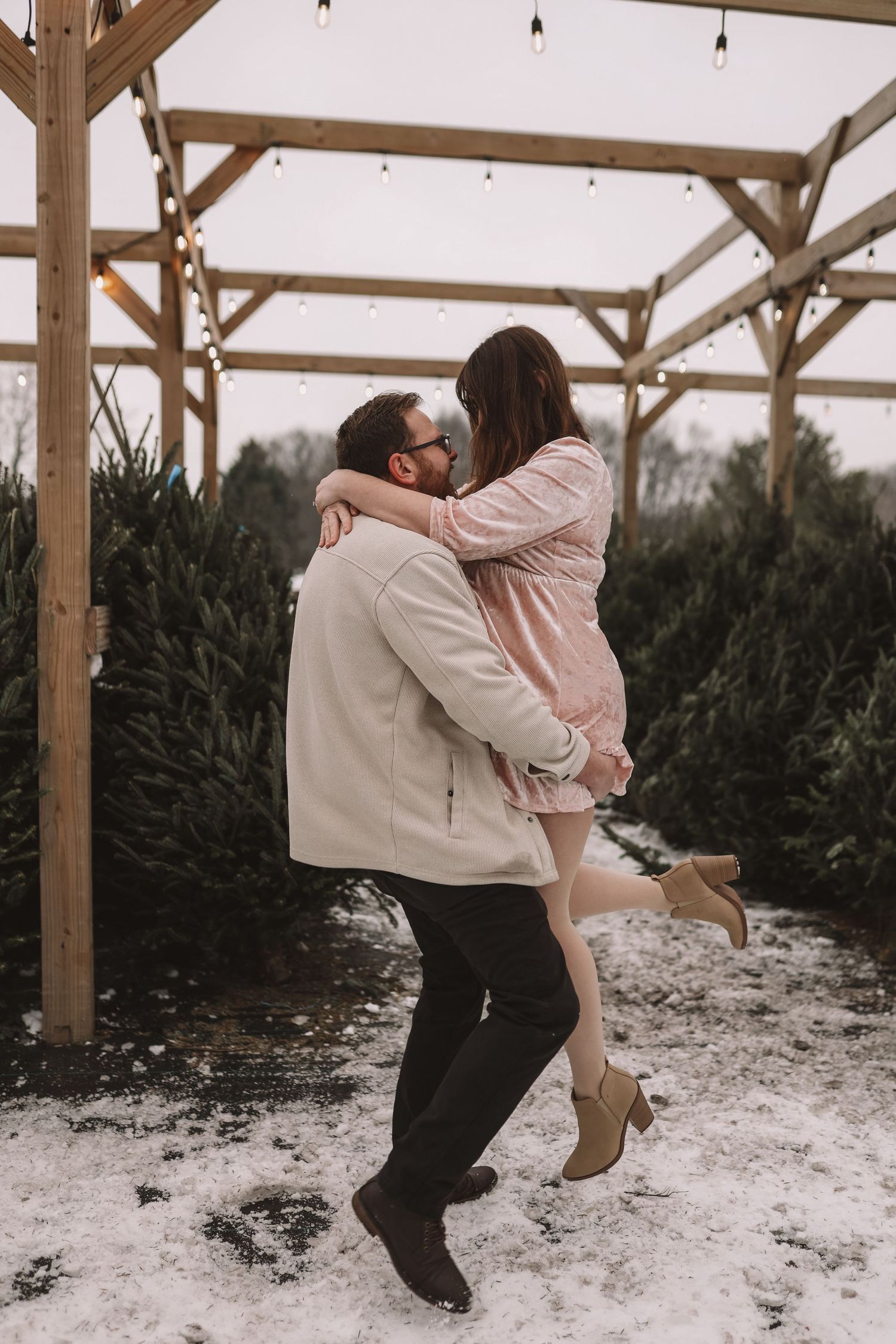 Couple sharing a romantic kiss at a snowy Christmas tree farm under a wooden pergola with string lights.