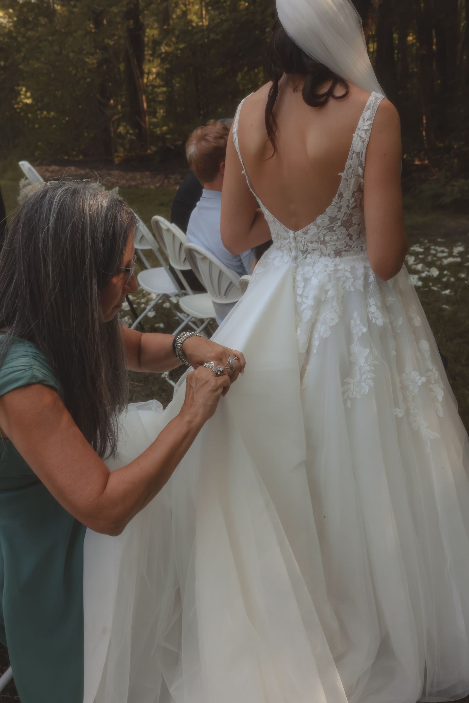 Woman adjusts bride's lace wedding gown train before outdoor woodland ceremony.