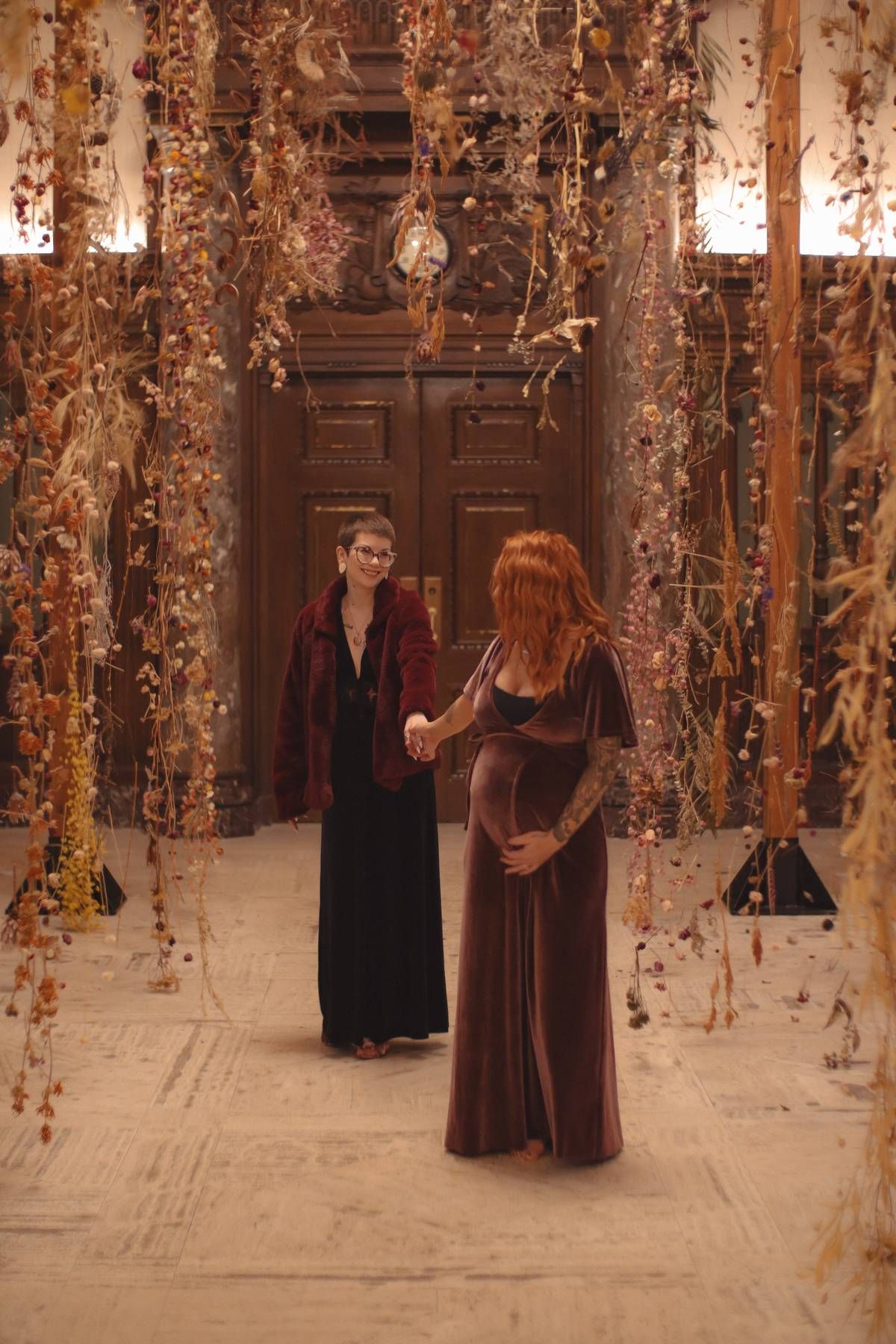 Two women hold hands in a floral installation with dried hanging flowers and a wooden door backdrop.