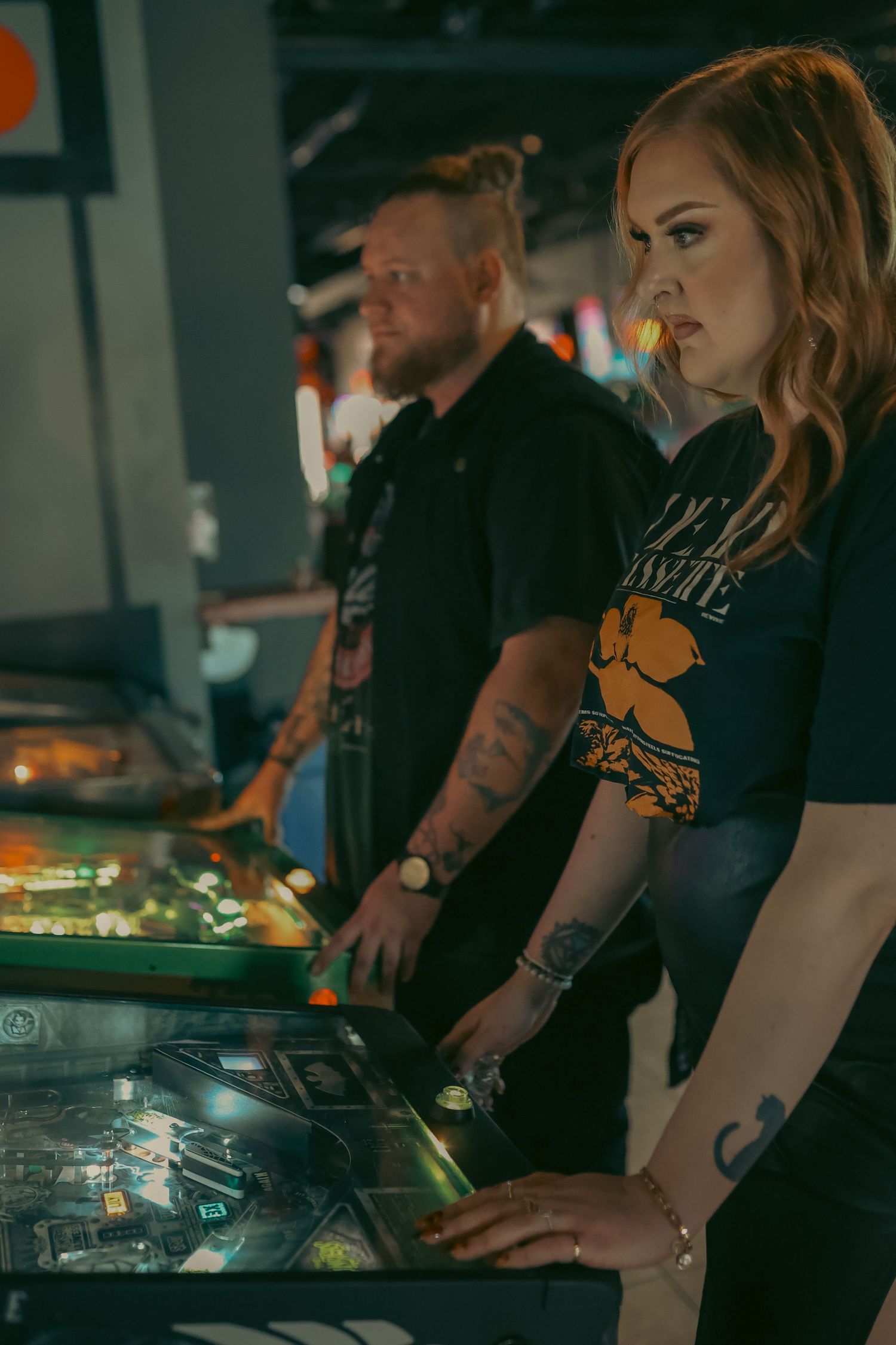 Two tattooed players focused on pinball machines in a dimly lit arcade, woman in blue tee, man in black shirt.