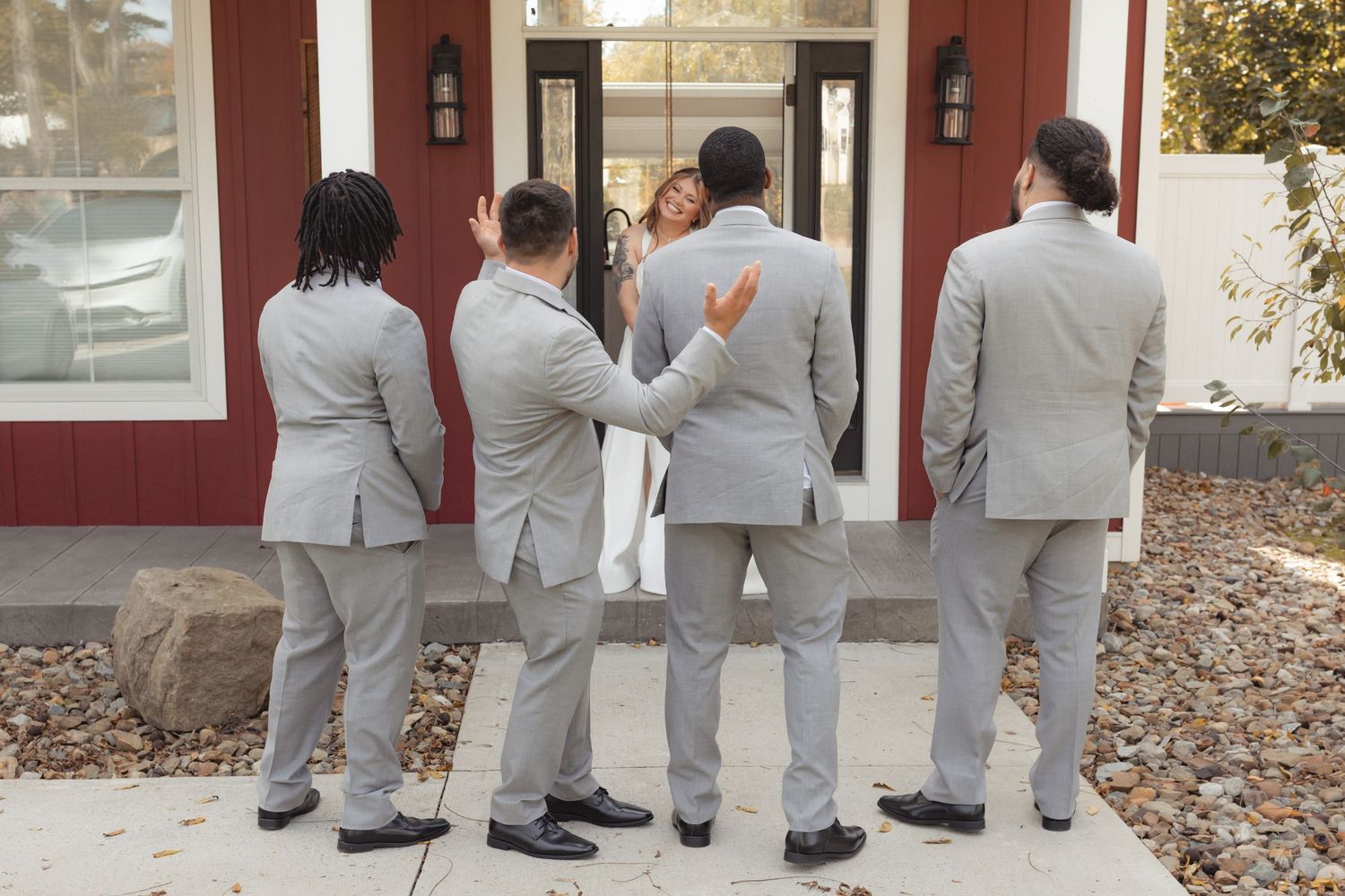 Groomsmen in gray suits stand outside a red building, reacting with surprise to a bride's reveal.
