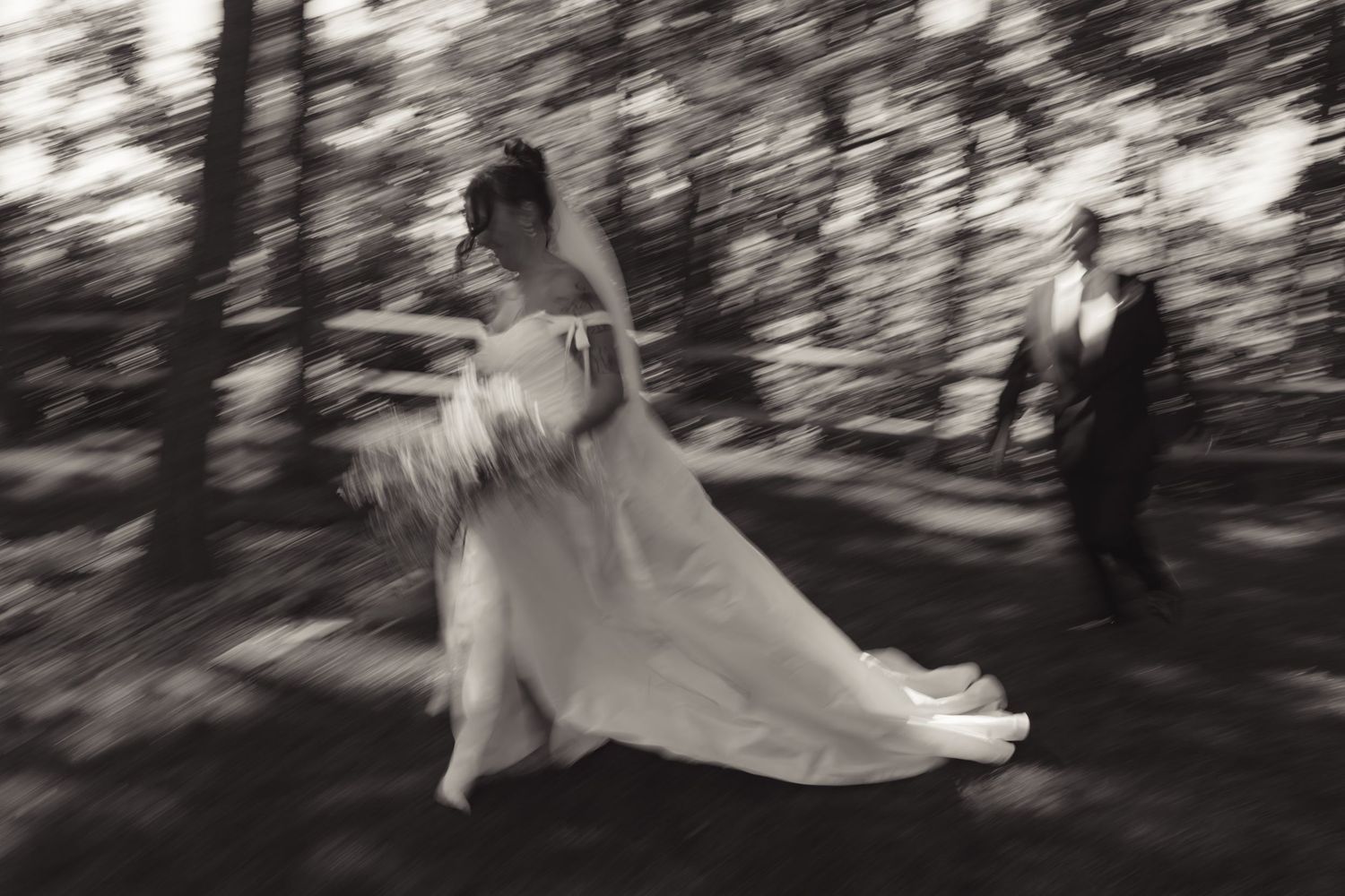 Black and white motion-blur photo of a bride in a flowing gown and groom walking through a wooded area.