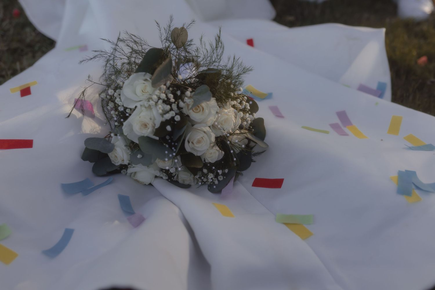 White rose bouquet with greenery resting on white fabric scattered with colorful confetti pieces outdoors.